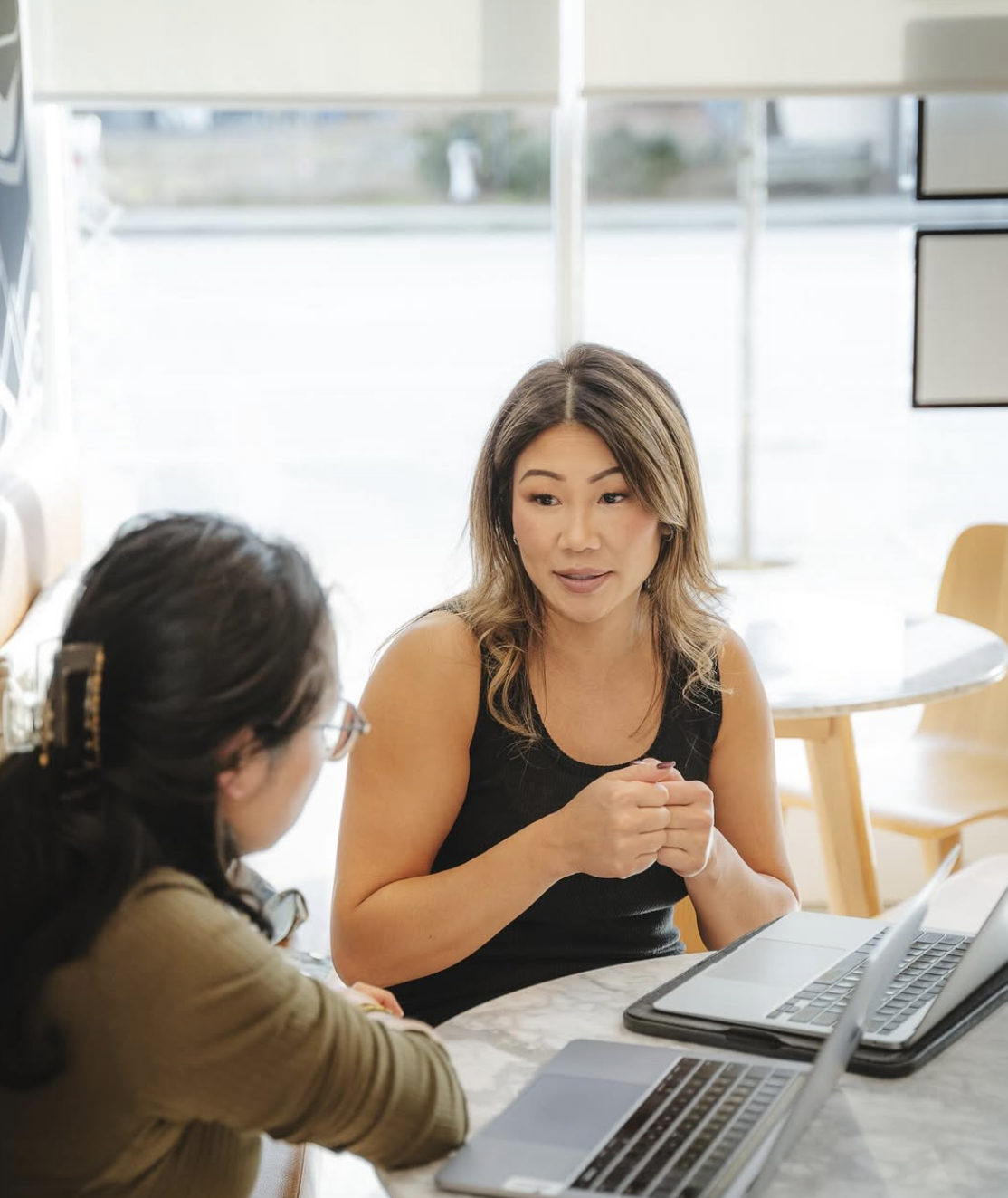 Two women having a conversation at a table in a bright indoor setting with laptops.