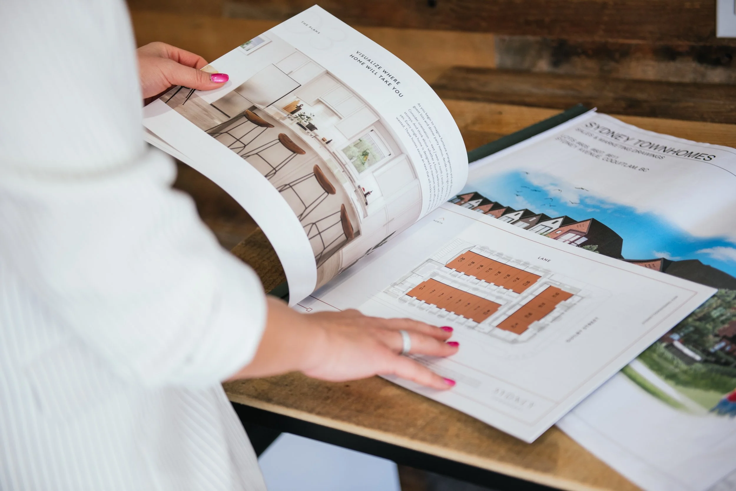 A person wearing a white shirt with bright pink nails holding open a real estate brochure displaying interior photos and floor plans of homes.