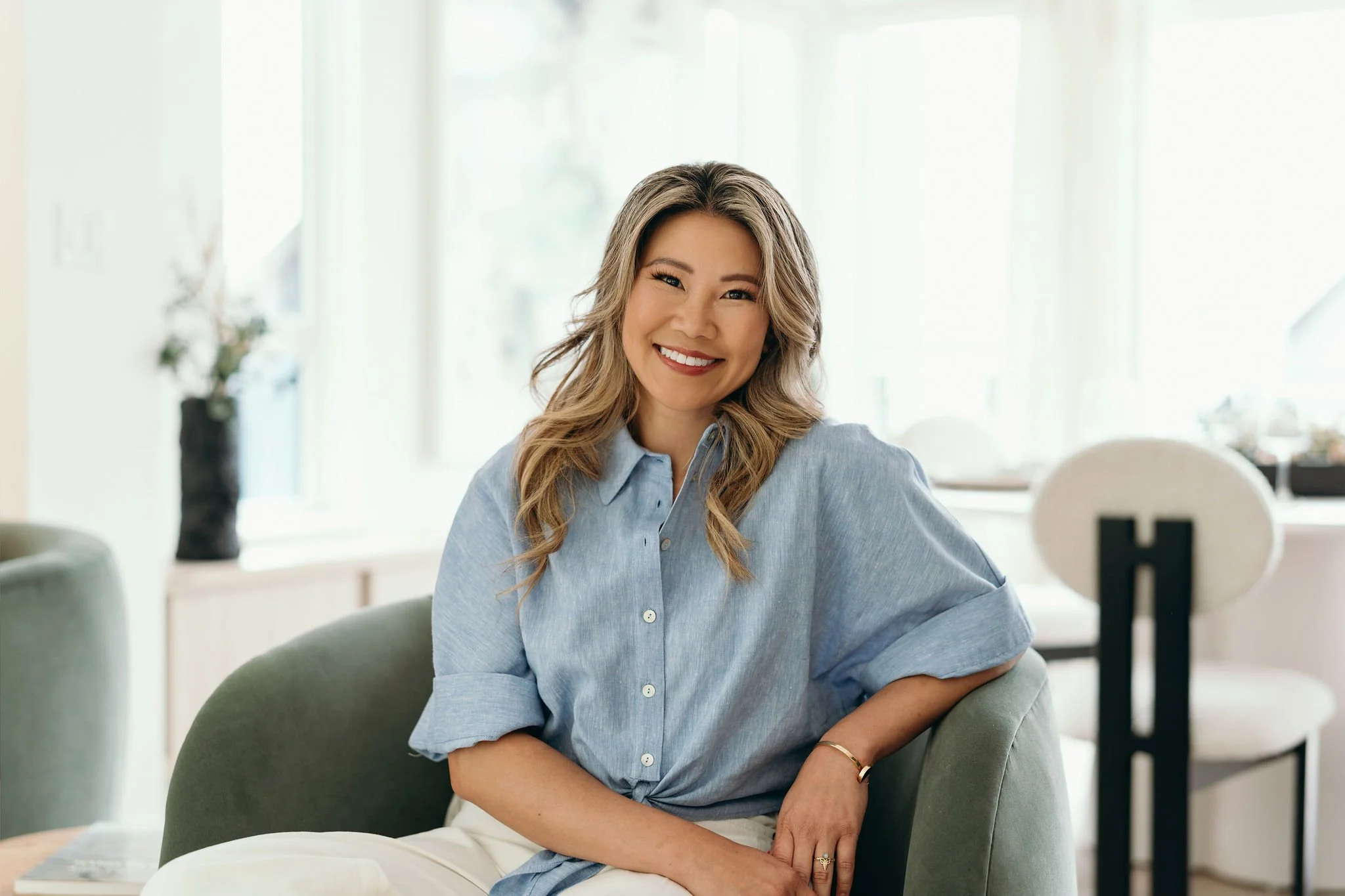 A woman with styled blonde hair, wearing a blue shirt, smiling and sitting on a green armchair in a bright room.