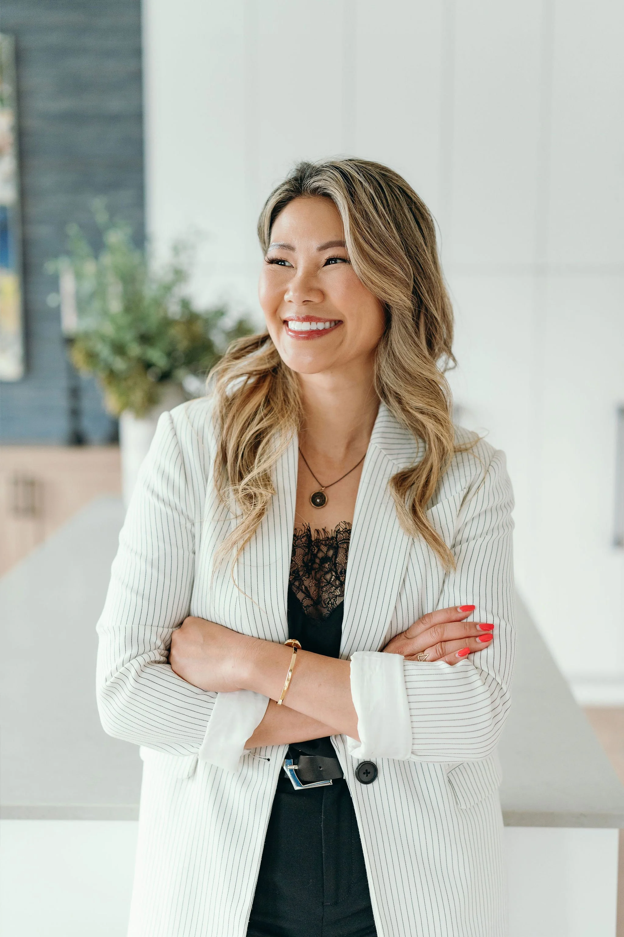 Smiling woman with wavy blonde hair, wearing a white pinstripe blazer, black lace top, gold jewelry, and red lipstick, standing with crossed arms in an office space with a blurred background.