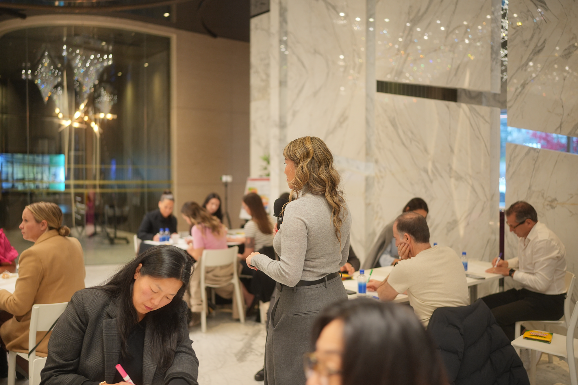 A woman with wavy blonde hair and a gray blazer speaking into a microphone during a meeting or conference. Several people are sitting at tables, some taking notes, in a modern, well-lit room with marble walls and decorative lighting.