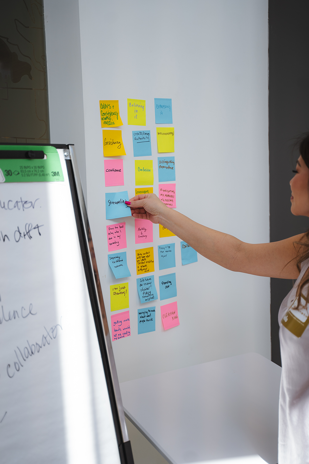 A woman sticks a blue sticky note on a wall filled with colorful sticky notes organized in columns for a planning or brainstorming session.