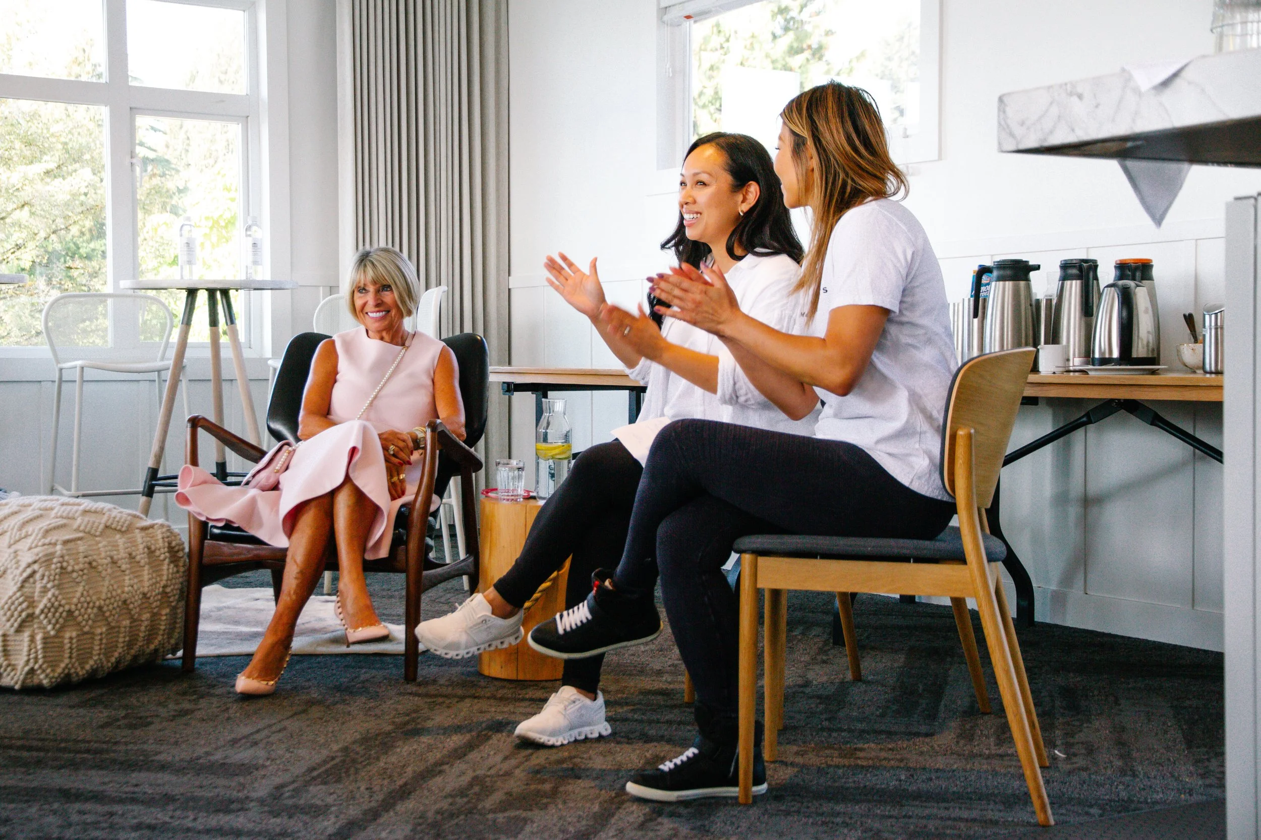 Three women are sitting and chatting in a bright, modern room with large windows. One woman in a pink dress is seated, smiling, while two women in casual white t-shirts are sitting on chairs, engaged in conversation. There are kitchen appliances and a table in the background.