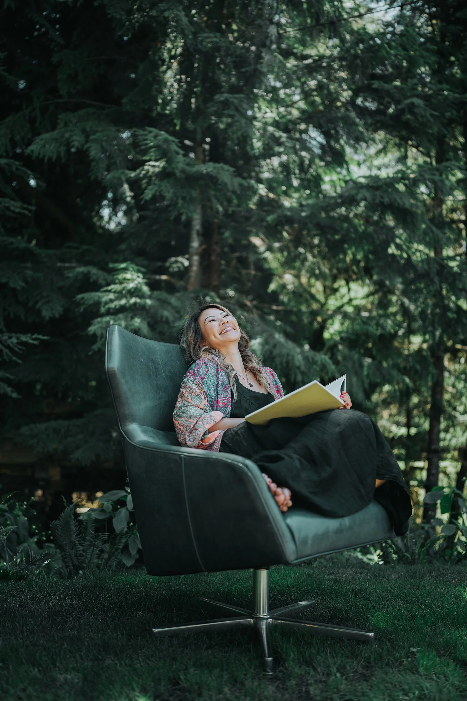 A woman sitting comfortably in a modern swivel chair outdoors, surrounded by lush green trees and plants, enjoying reading a book and smiling.