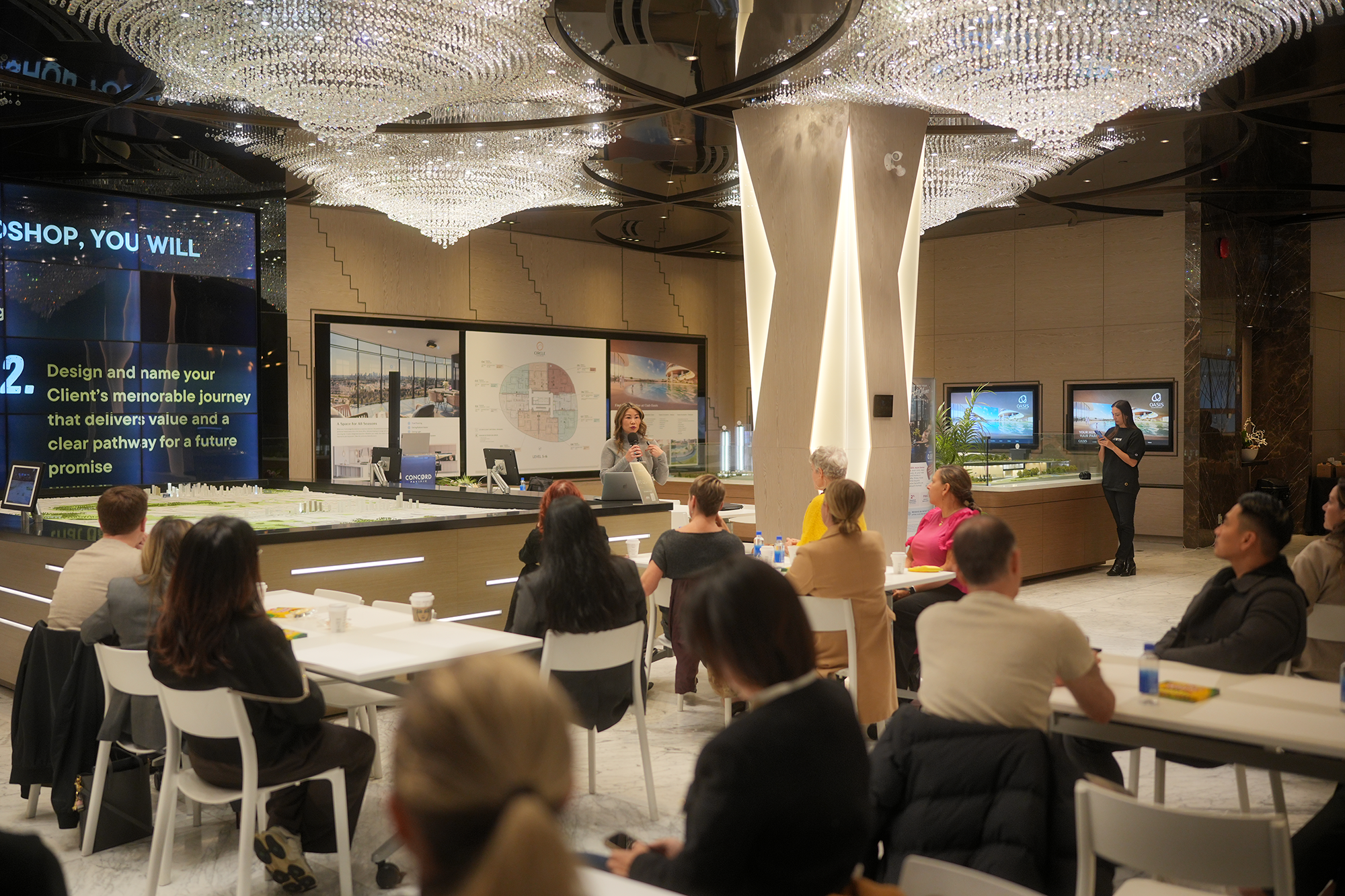 A woman is speaking into a microphone at a presentation in a brightly lit lobby with chandeliers. The audience sits at white tables, and television screens display promotional images. The room also features large display screens and posters related to real estate or architecture.