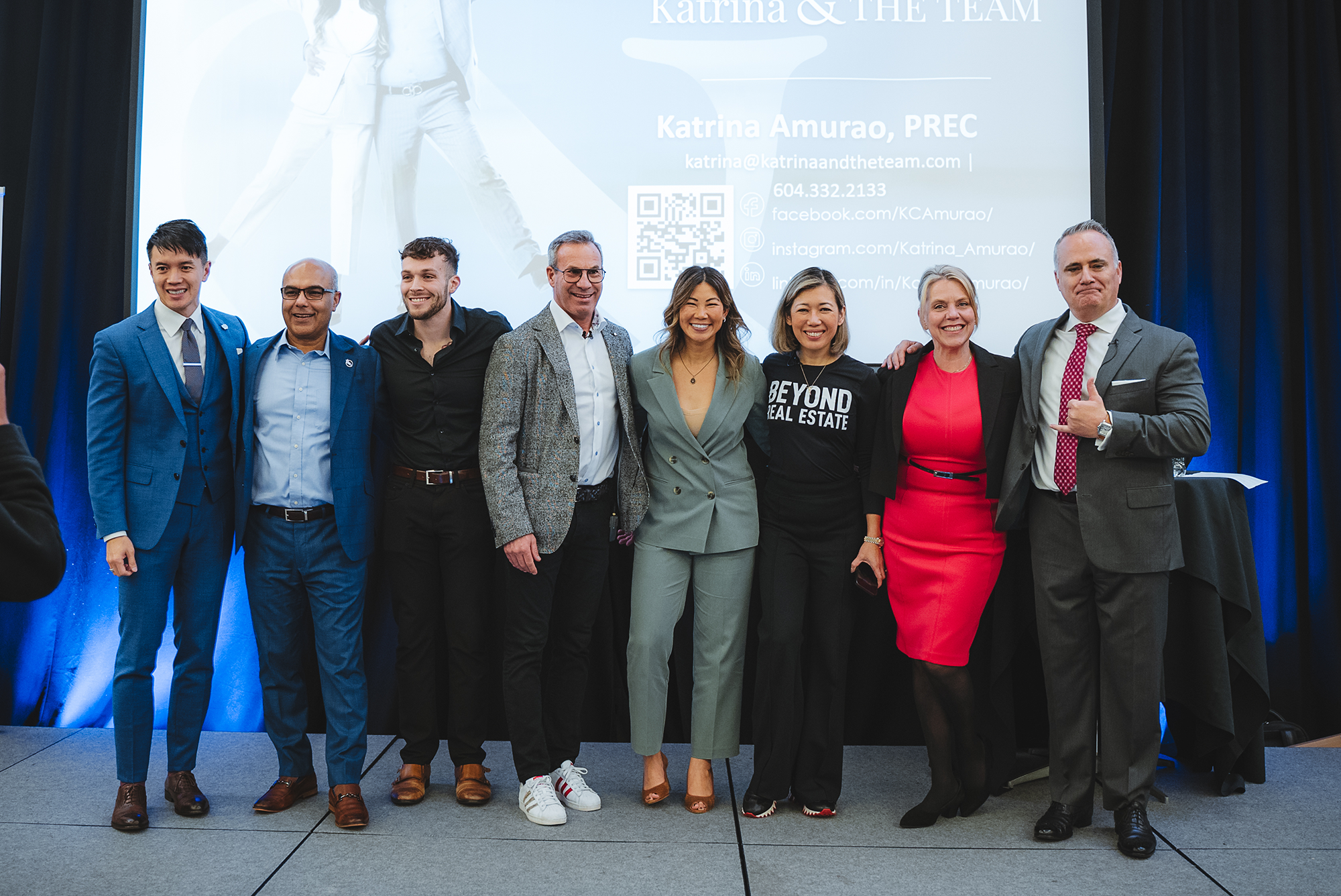 A group of eight diverse professionals standing together indoors, smiling for a photo, in front of a presentation screen displaying contact information for Katrina Amurao, including email, phone number, and social media links.