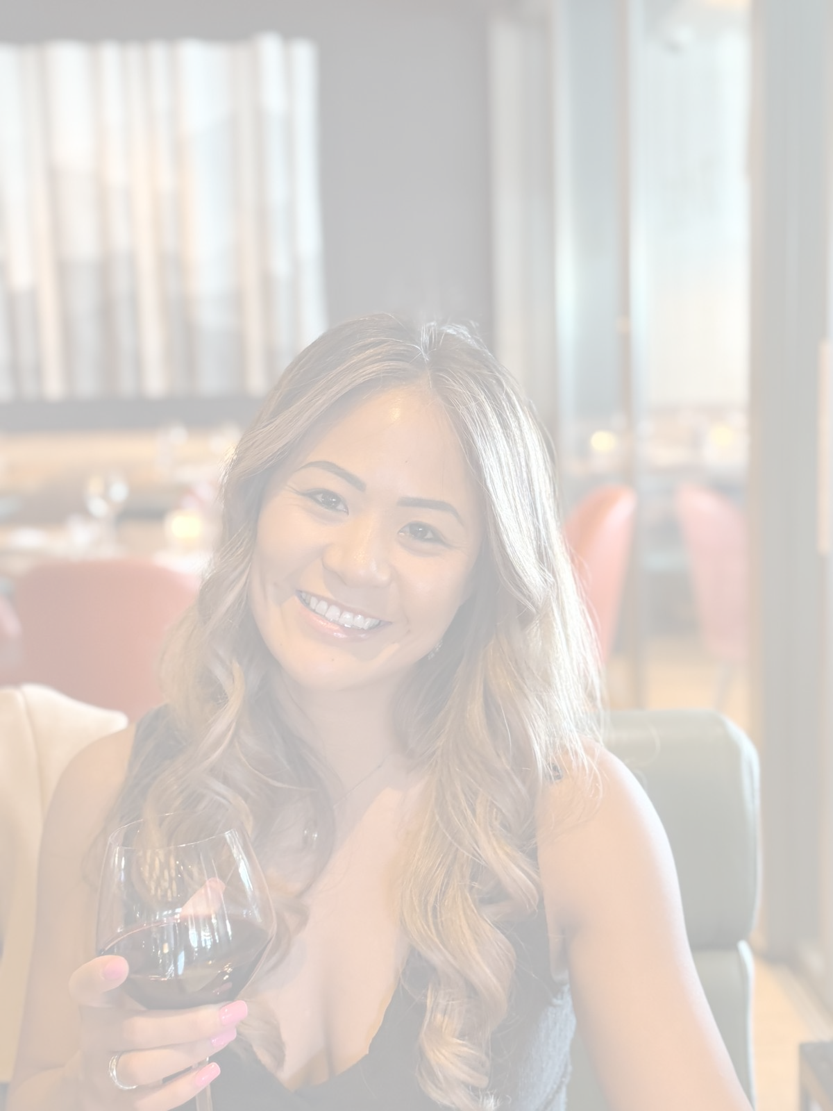 A woman with long wavy hair smiling and holding a glass of red wine, sitting in a restaurant with natural light coming through curtains in the background.