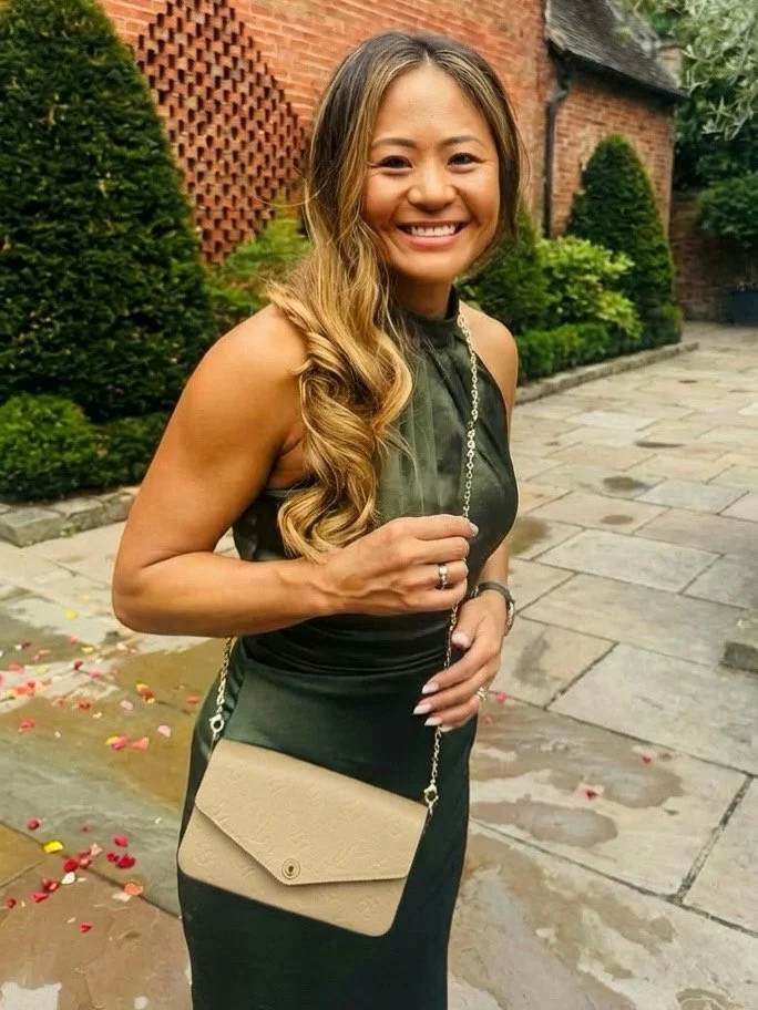 A woman smiling outdoors, wearing a black dress with a leopard print strap, holding a beige purse with a chain strap, standing on a stone pathway with flowers and greenery in the background.