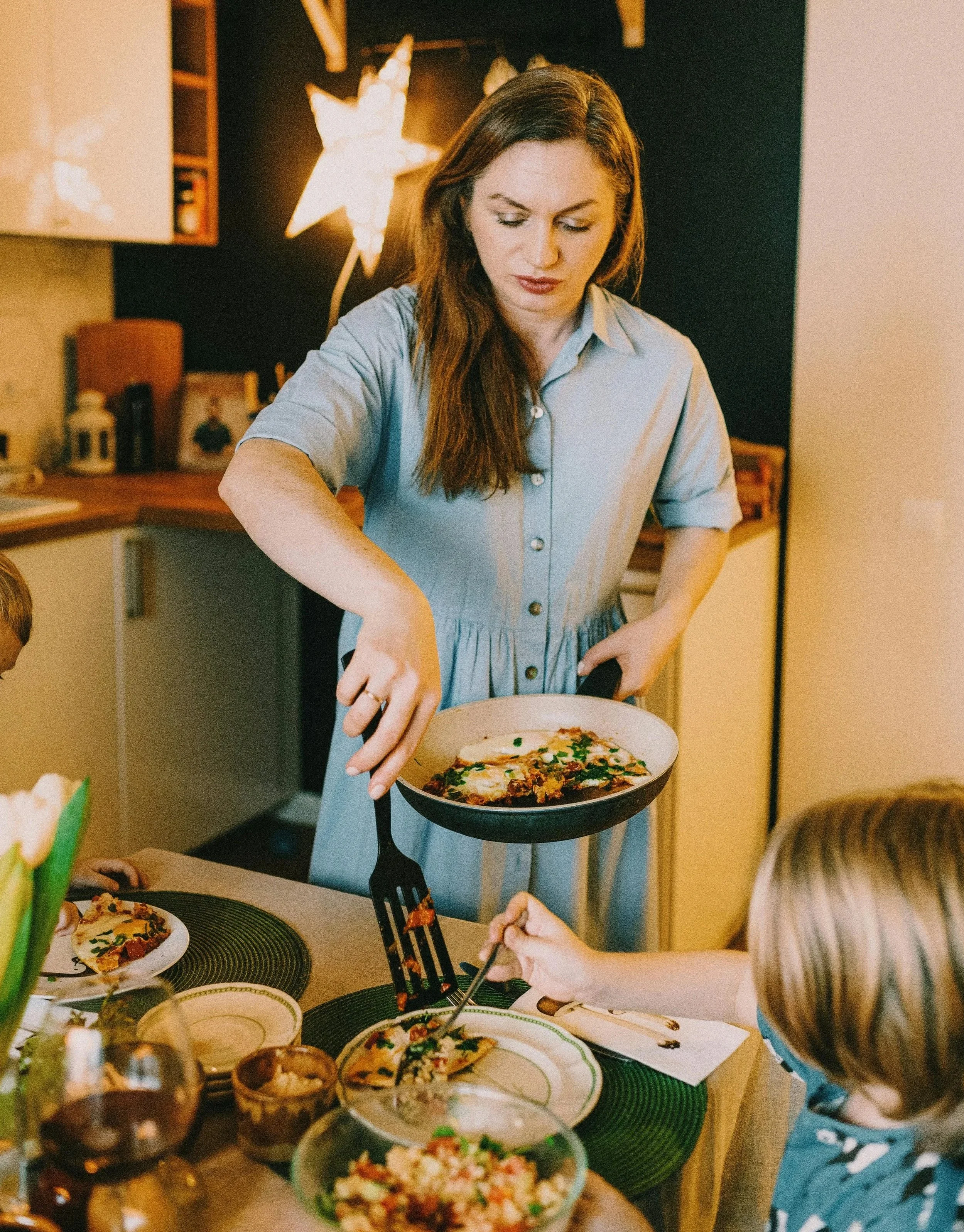 mother wearing a blue collared shirt cooking breakfast with a pan in a kitchen with her child