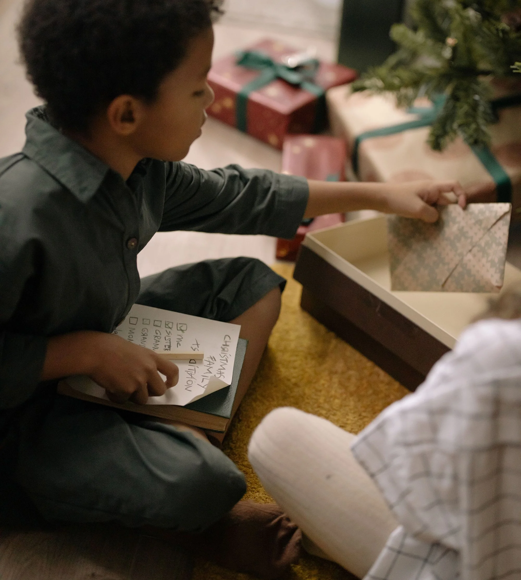 child with christmas wishlist sitting cross-legged by a tree reaching for a wrapped present