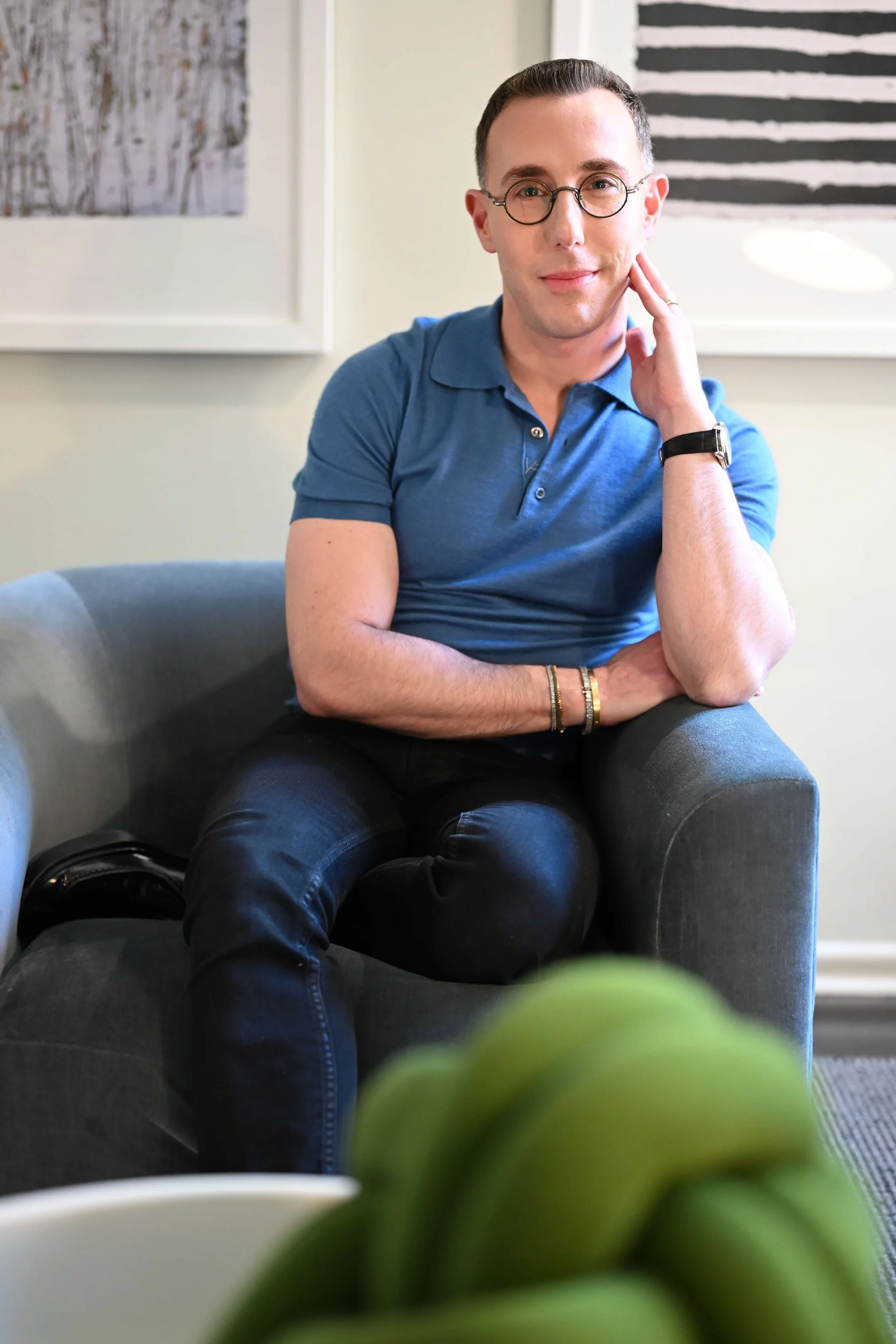 Dr Josh Mirmelli - A man sitting on a gray chair, wearing glasses and a blue polo shirt, with a short hairstyle and a watch on his left wrist, resting his face on his hand in a room with artwork on the wall behind him.