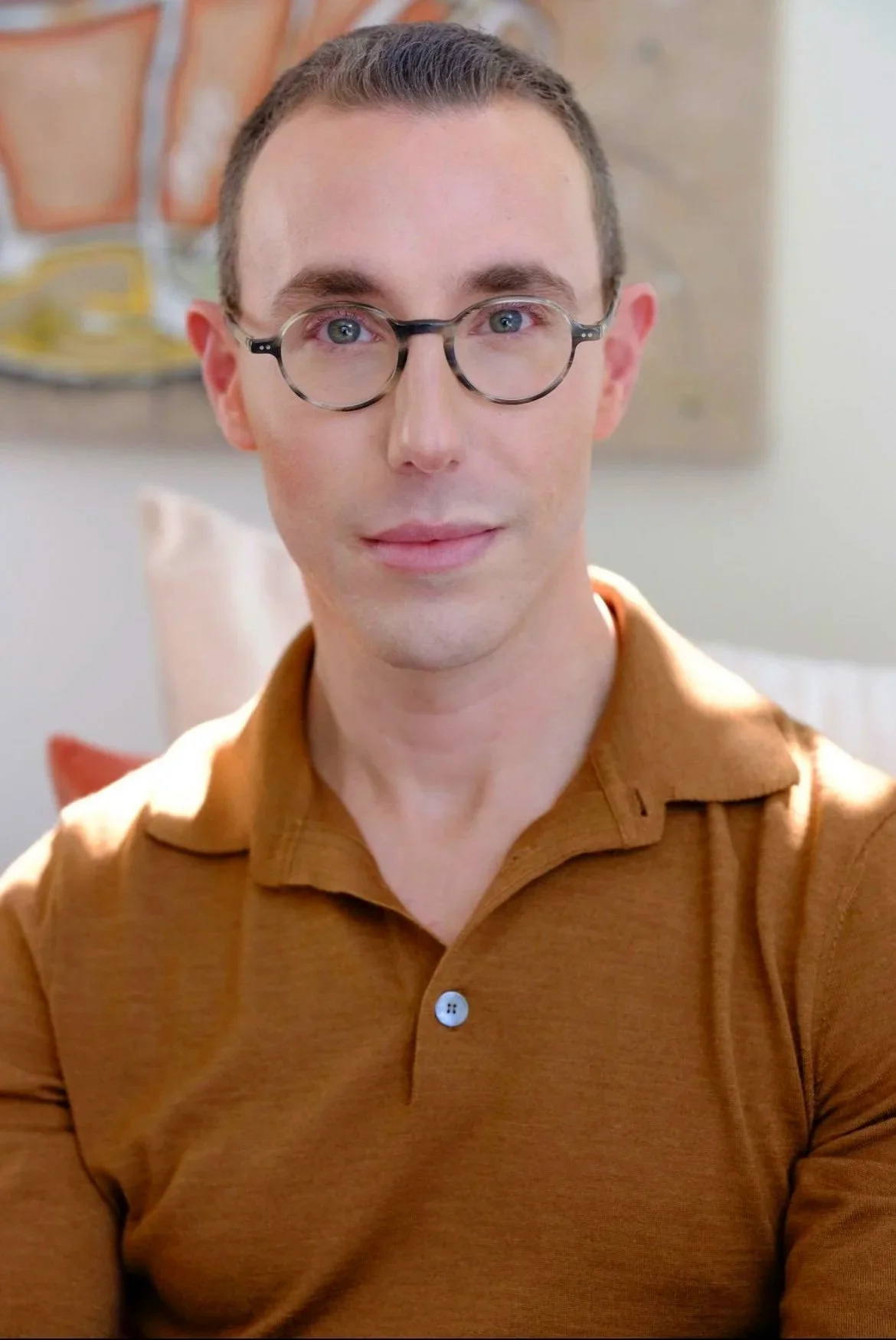 Dr Josh Mirmelli a man with light skin, short brown hair, and round glasses wearing a brown collared shirt, looking directly at the camera with a slight smile. In the background, there is abstract wall art and a white pillow.