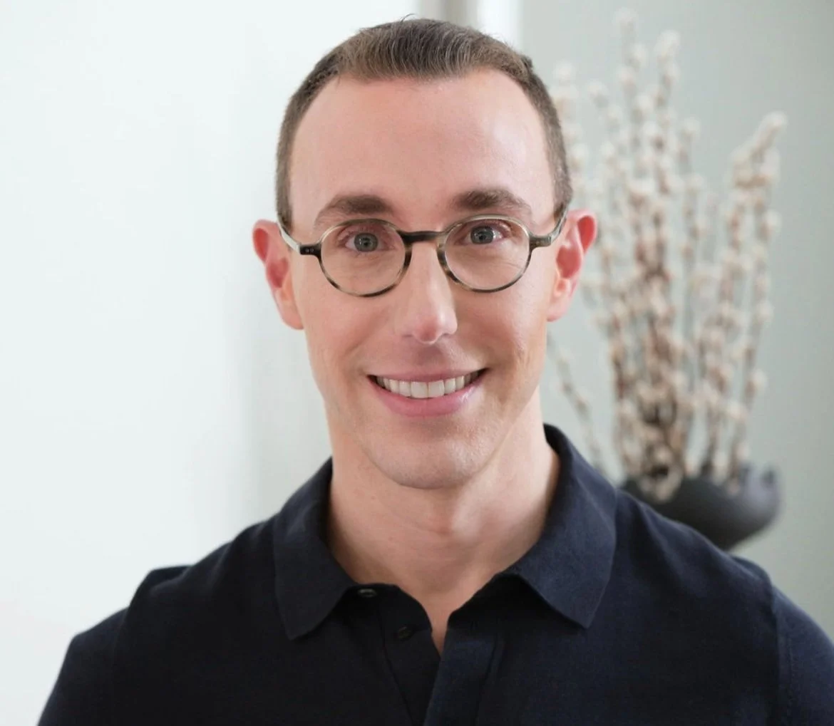 A smiling man with short brown hair, glasses, wearing a black collared shirt, standing in front of a white wall with a blurred plant in the background.