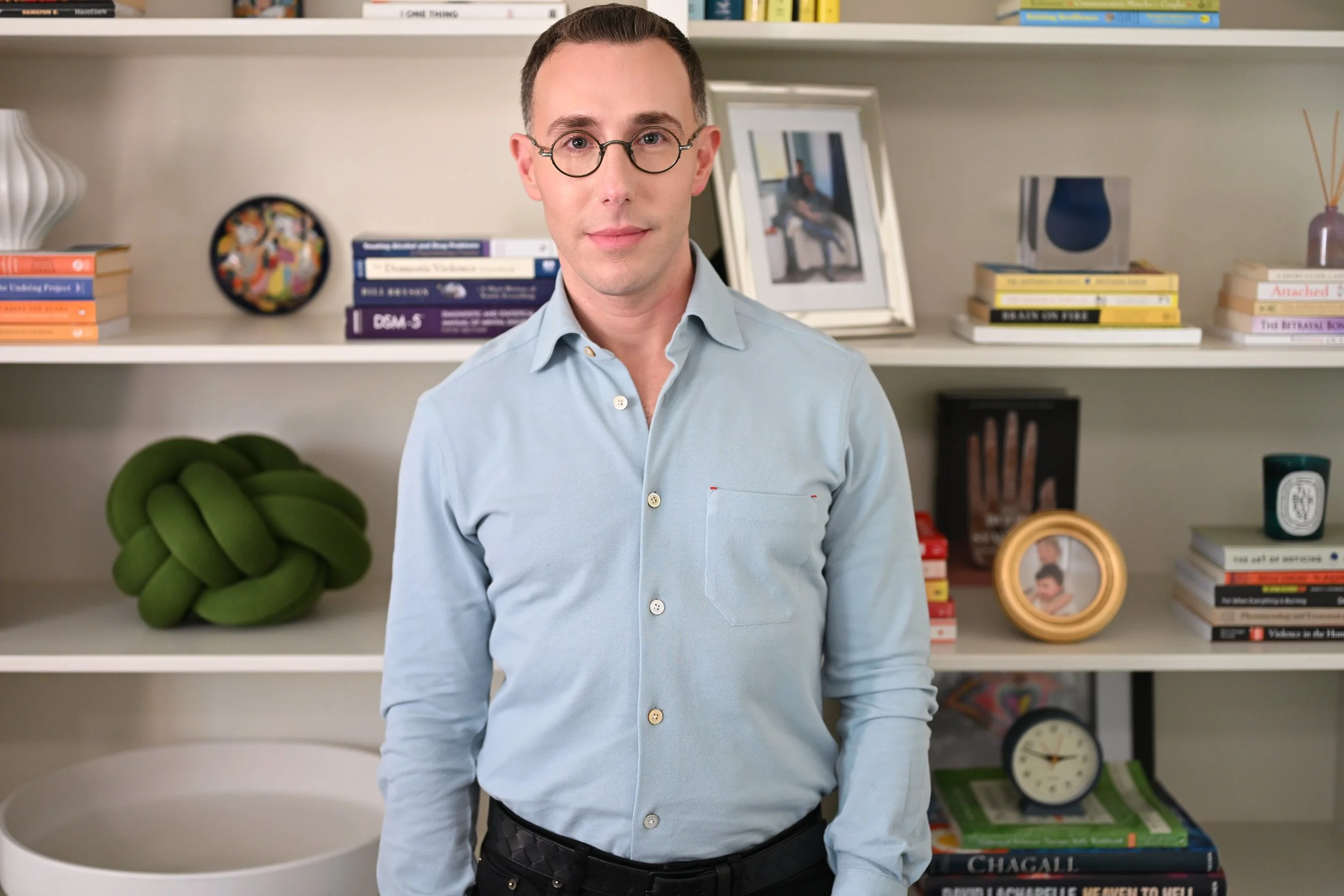 A man with glasses in a light blue shirt stands in front of a bookshelf. The bookshelf contains books, framed photos, a round decorative item, a clock, and other objects.