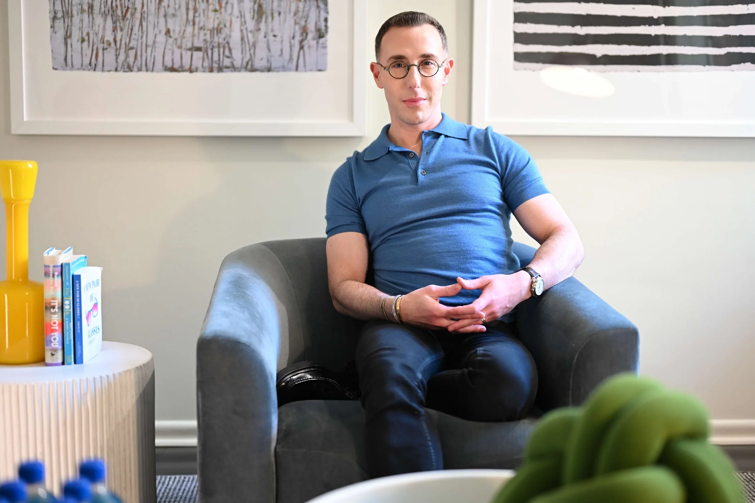 Dr Josh Mirmelli - a man with glasses and short hair, wearing a blue polo shirt and dark pants, sitting comfortably in a gray armchair in a modern room with framed art on the wall.