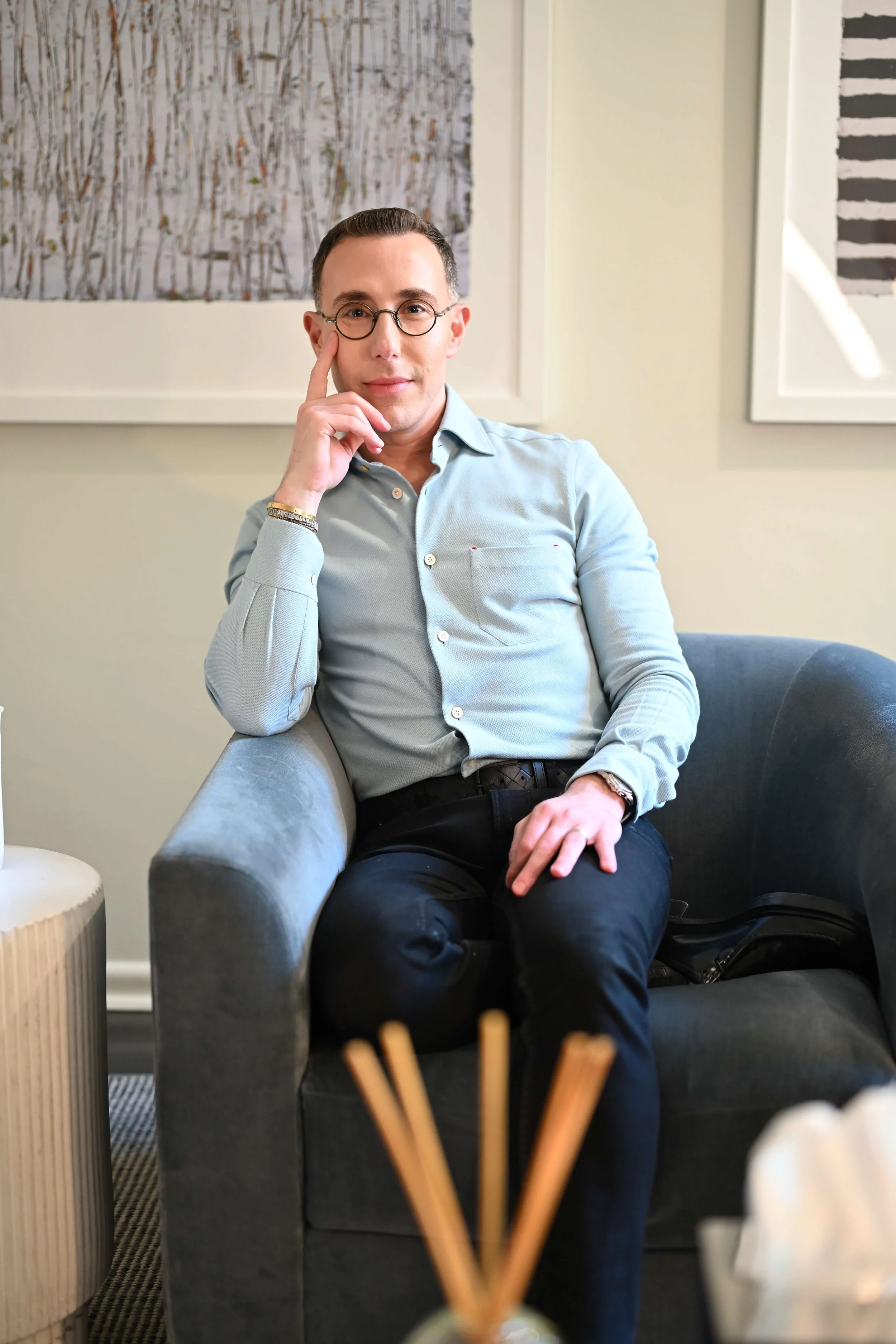 Dr Josh Mirmelli - a man with glasses, wearing a light blue button-up shirt, sitting on a gray armchair in an indoor setting, with framed artwork on the wall behind him and decorative objects in the foreground.