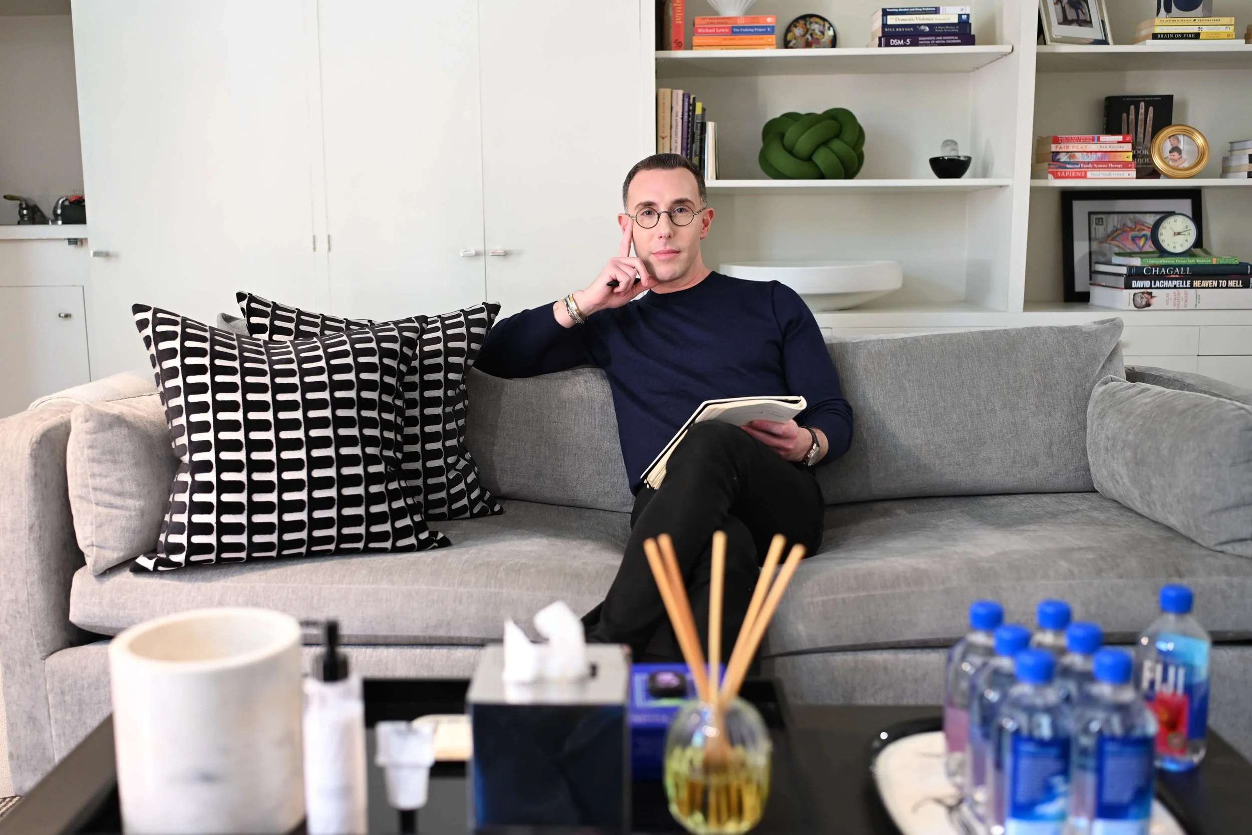 Dr Josh Mirmelli - A man sitting on a gray couch with patterned pillows, holding a book, in a living room with shelves filled with books and decorative items in the background.