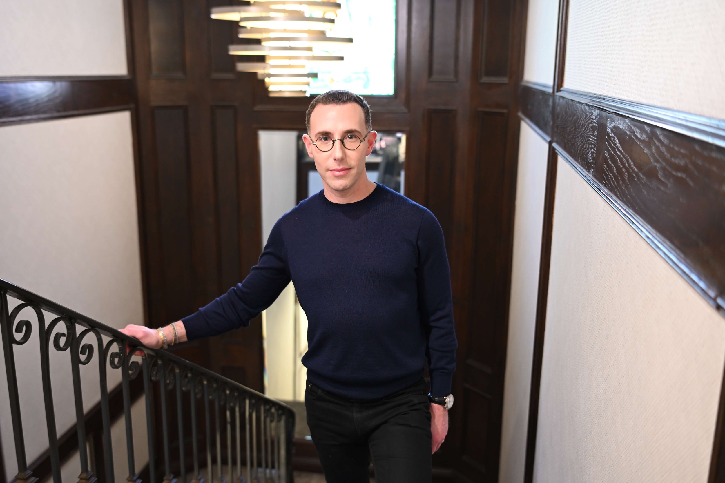Dr Josh Mirmelli - A man with glasses wearing a navy sweater and black pants standing by a staircase in a hallway with dark wood paneling.