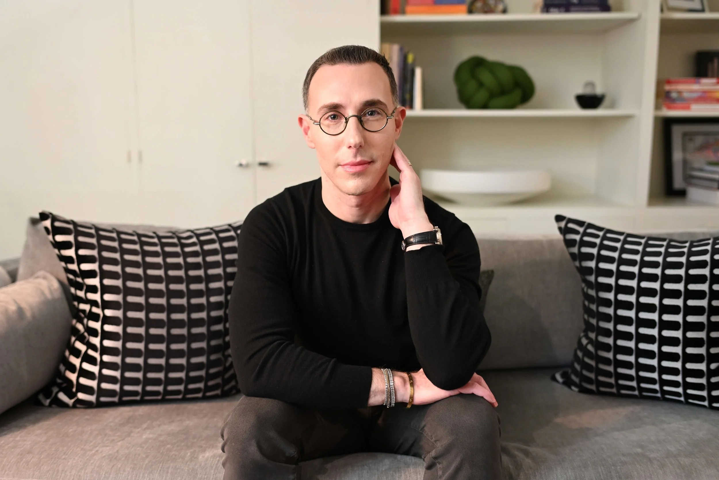 Dr Josh Mirmelli - A man with glasses and short dark hair sitting on a gray sofa with patterned pillows, in front of white shelves filled with books and decorative objects.