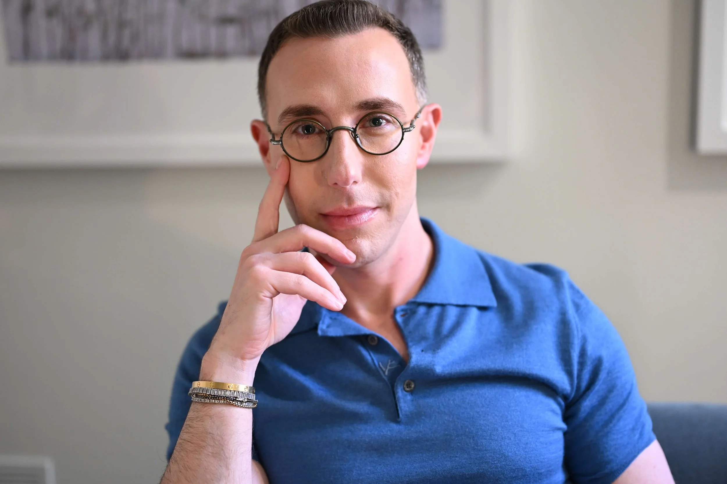 Dr Josh Mirmelli - A man wearing glasses and a blue polo shirt, sitting indoors with a neutral background, resting his face on his left hand.