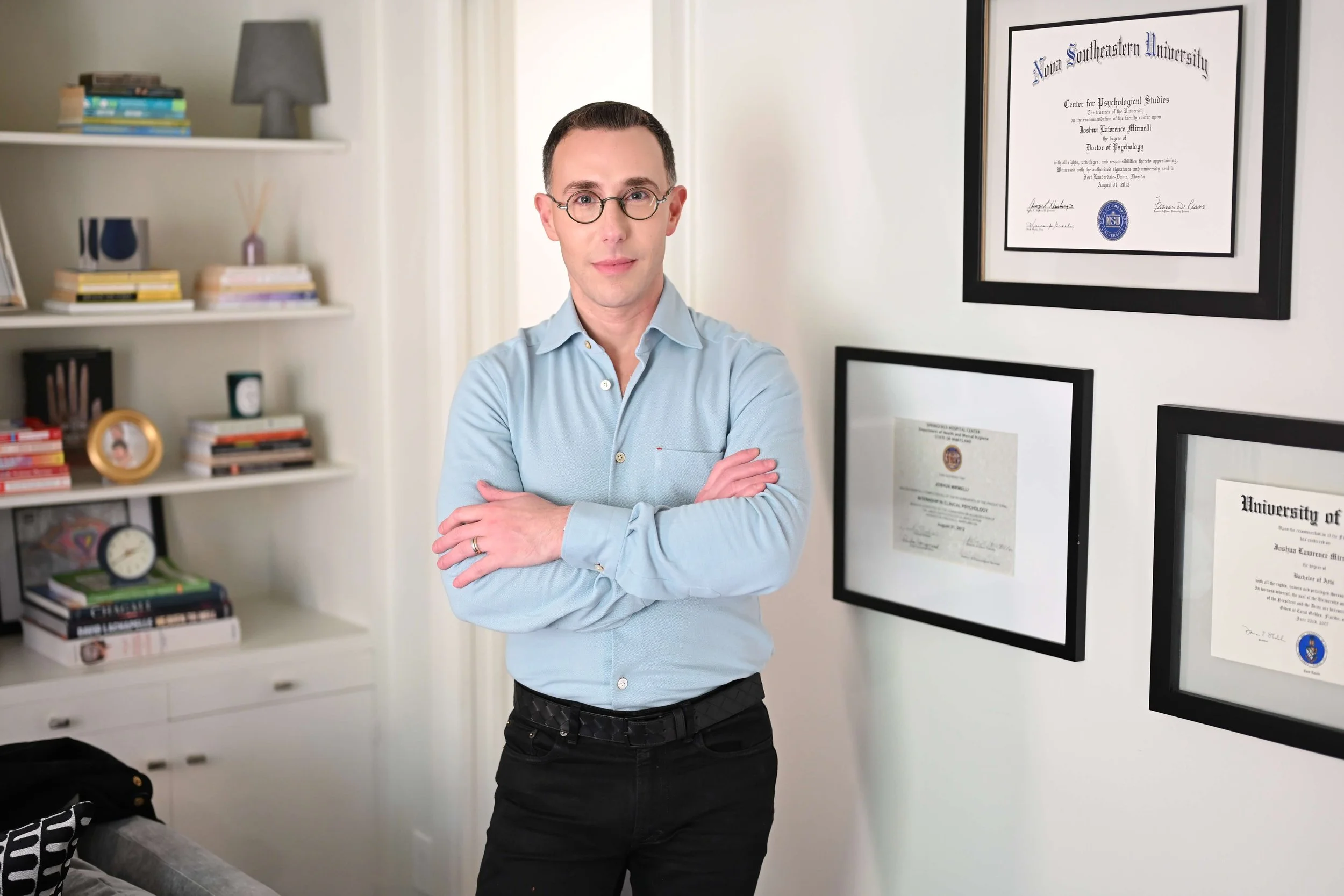 Dr Josh Mirmelli  - A man with glasses and a light blue dress shirt standing with arms crossed in a room with framed diplomas and a bookshelf.
