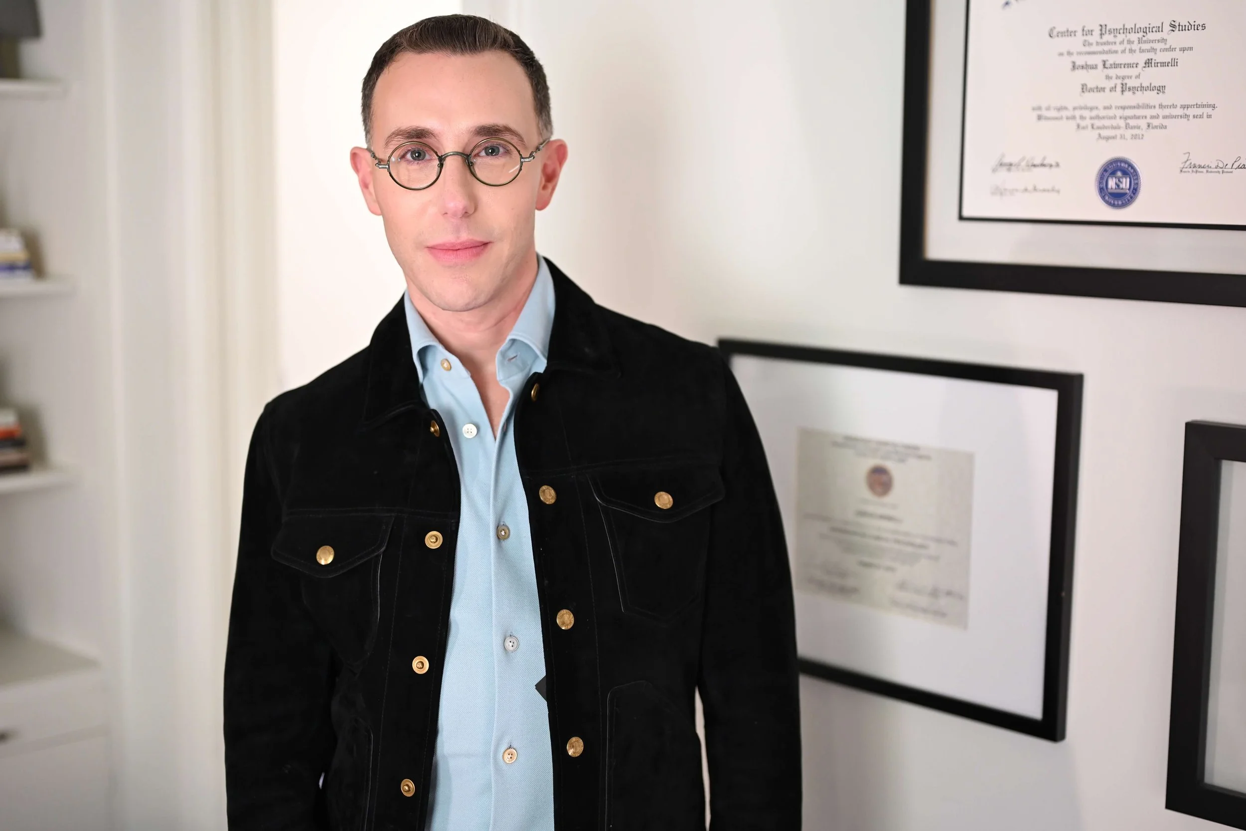 Dr Josh Mirmelli - A man with dark hair, wearing glasses, a light blue shirt, and a black jacket, standing indoors with framed certificates and papers on the wall behind him.