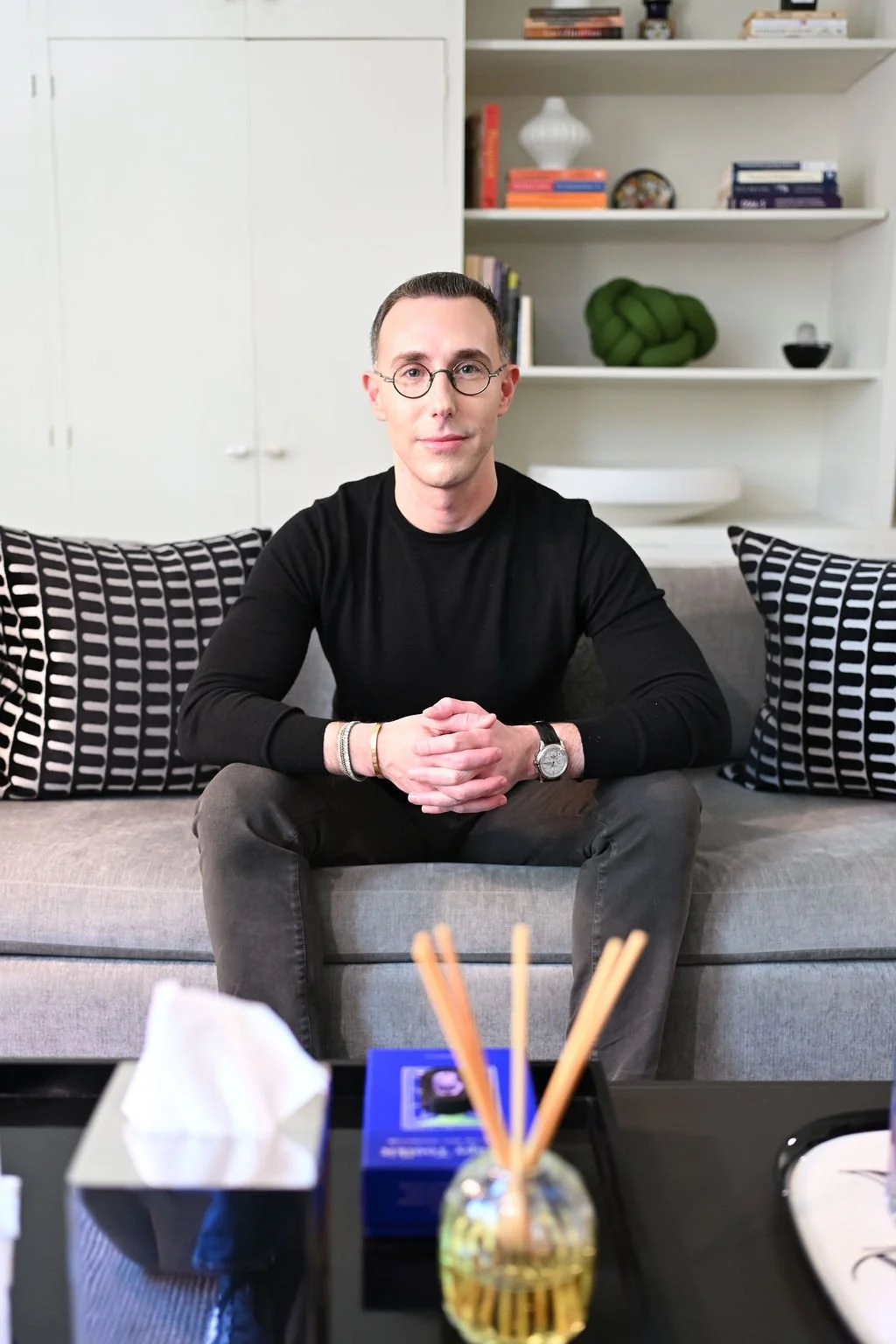 Dr Josh Mirmelli - A man sitting on a gray sofa with black and white patterned pillows, in a living room with a white bookshelf and decor in the background.