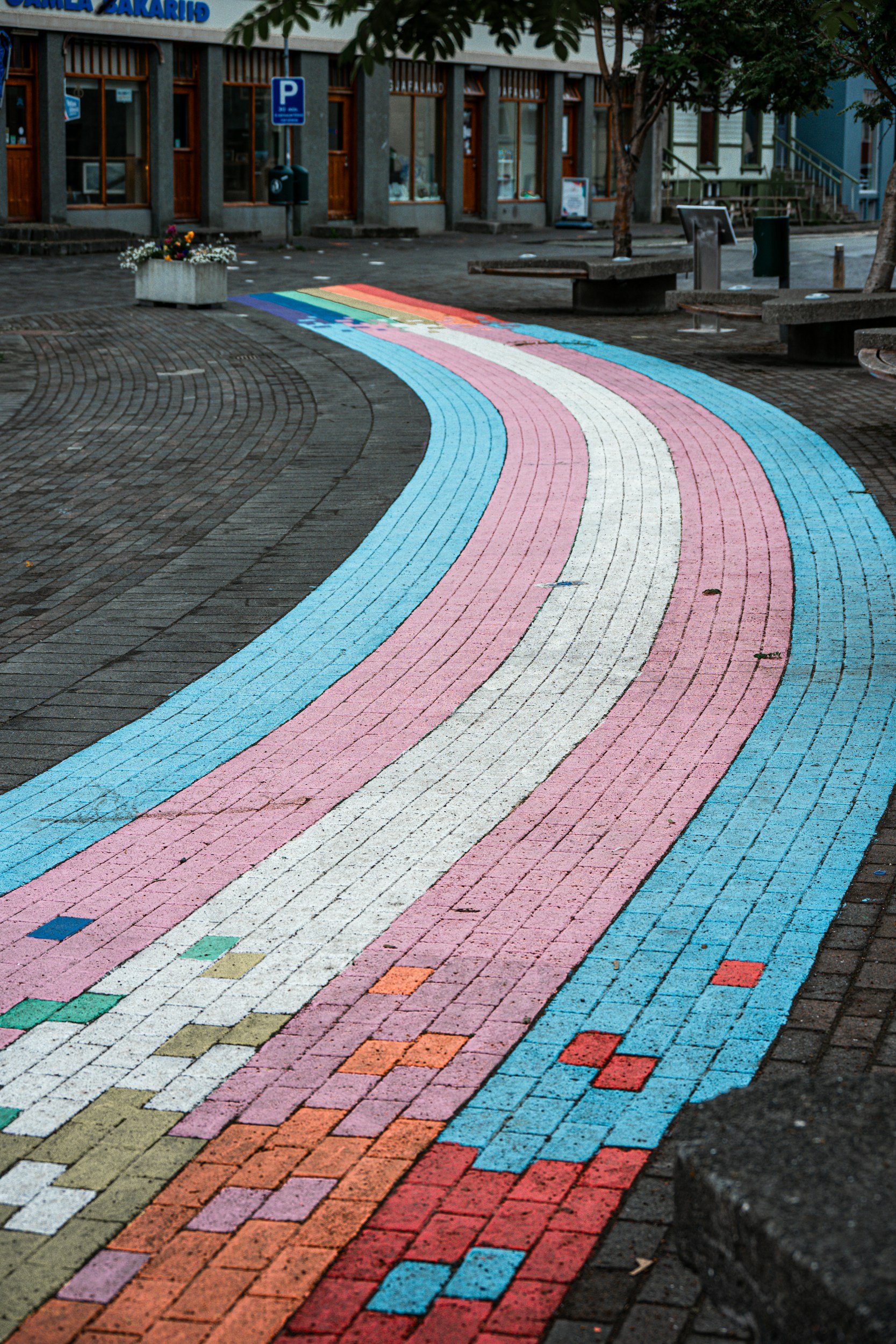 Colorful rainbow crosswalk painted on a city sidewalk with benches, trees, and storefronts in the background.