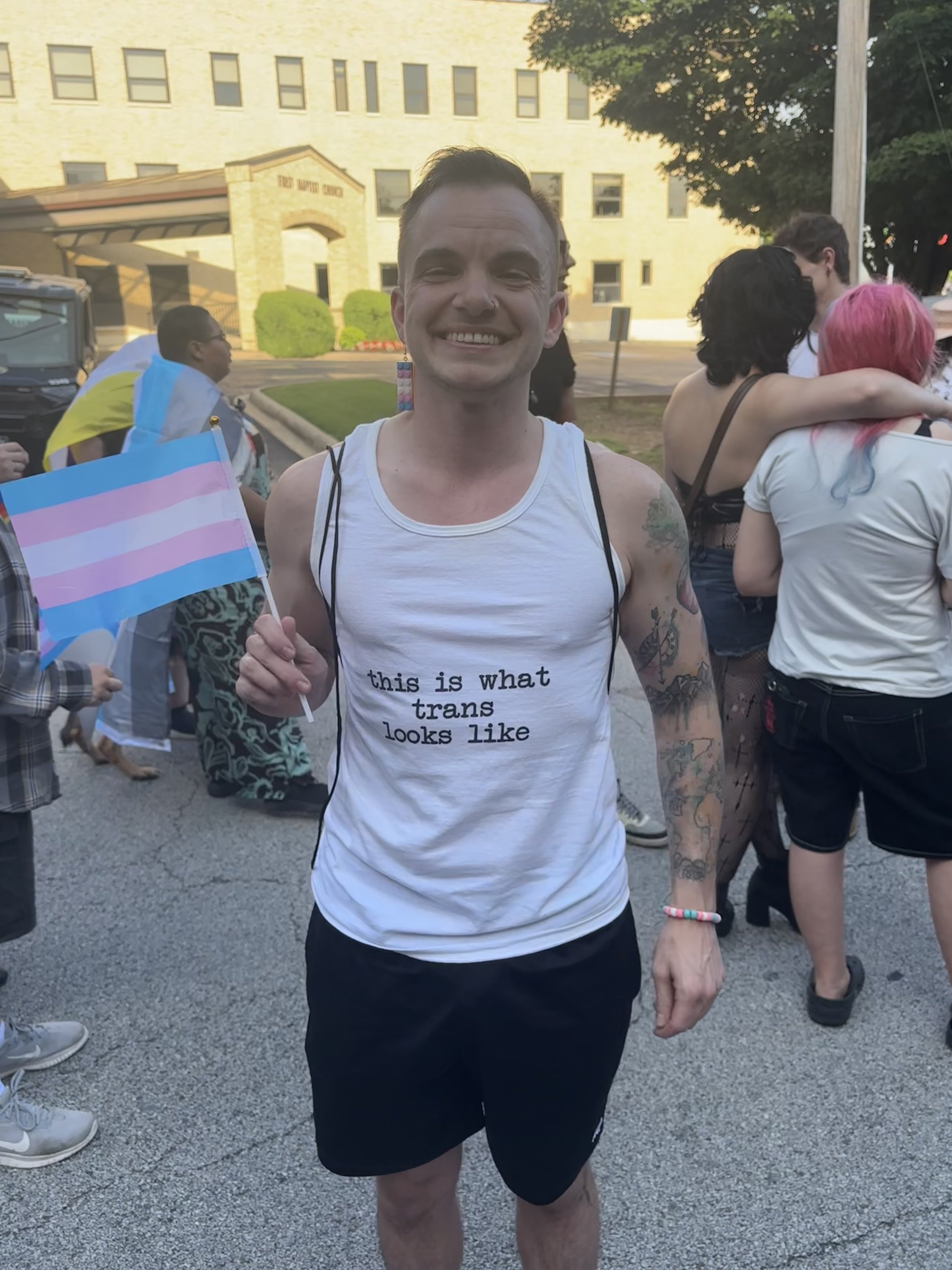 A smiling man holding a transgender pride flag at an outdoor event with other people in the background.
