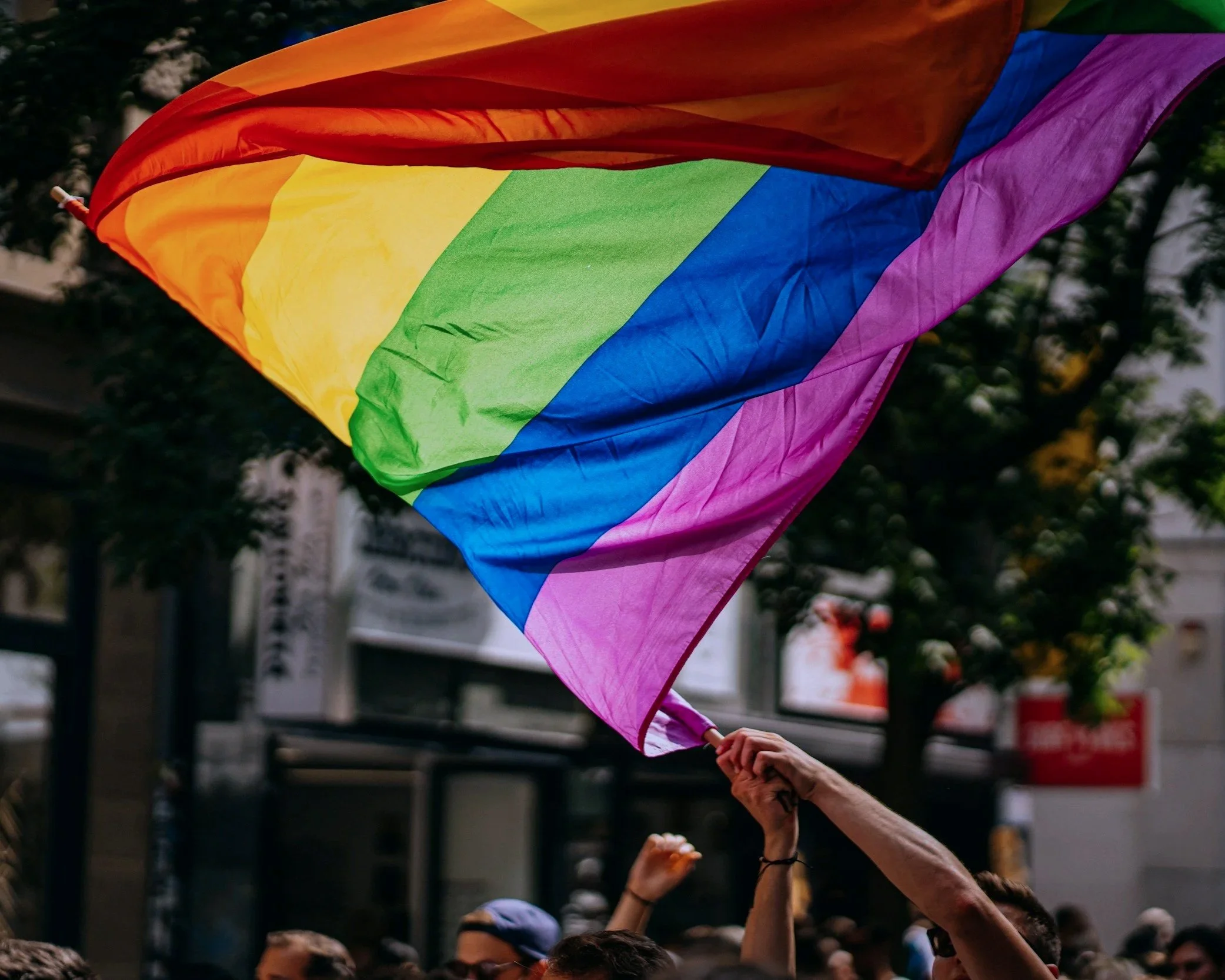 Rainbow pride flag being held up by multiple people during a parade or rally.