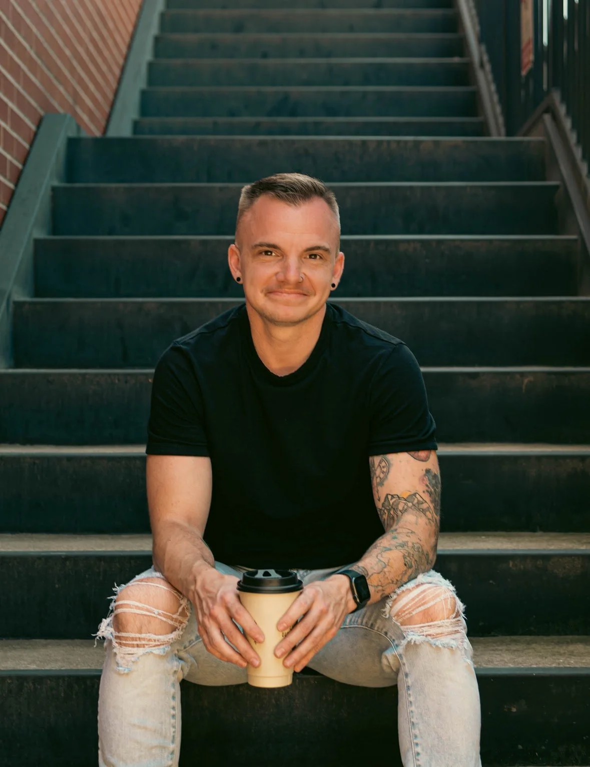 A young man sitting on outdoor black stairs, holding a to-go coffee cup, smiling at the camera, wearing a black t-shirt, ripped jeans, and tattoos on his left arm.