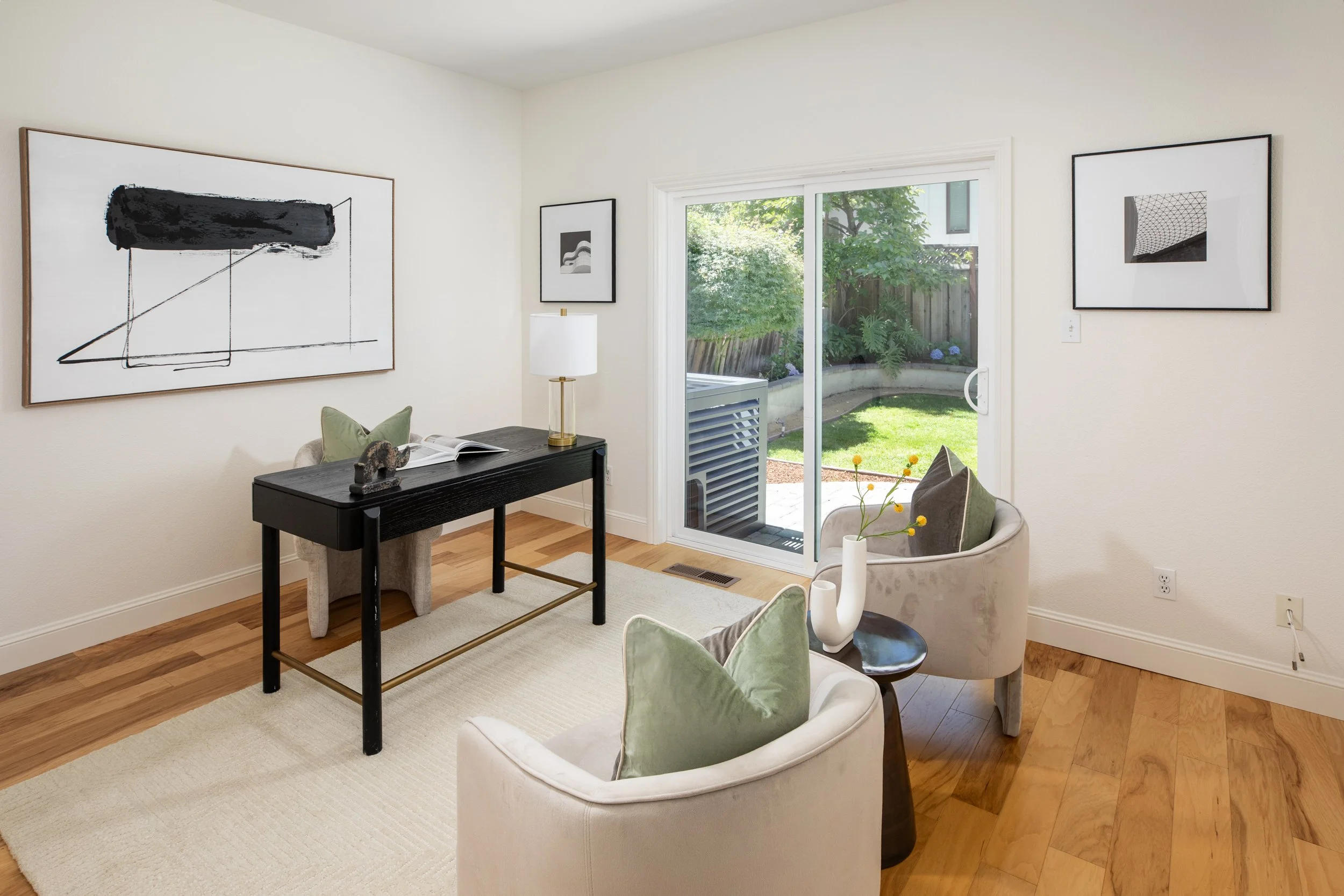 A cozy living room with a small black table, striped beige and gray armchair, and some green and gray pillows. Decor includes framed abstract art on white walls, a white lamp with a gold base, and a white vase with yellow flowers. Large sliding glass