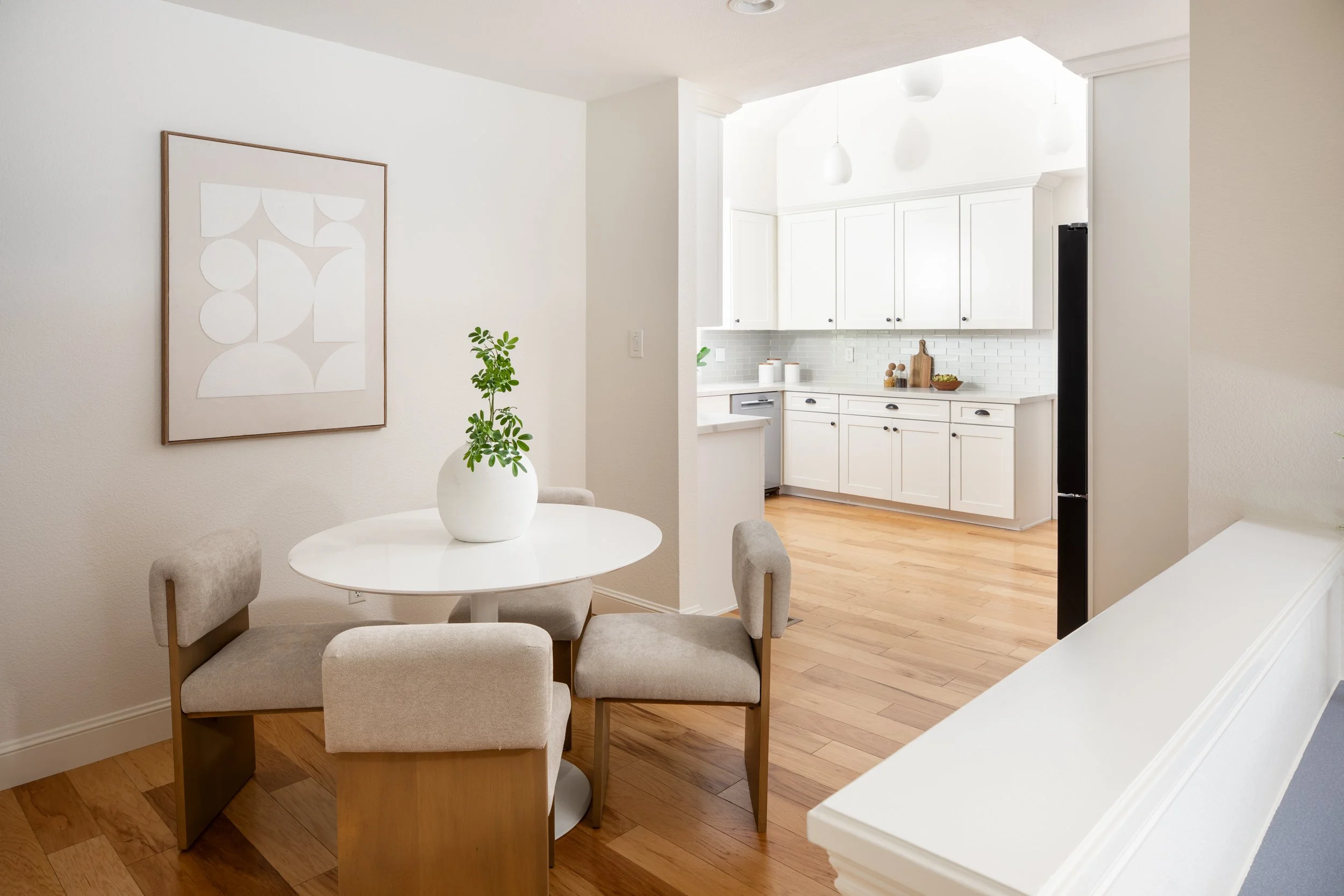 A dining area with a white round table, four cushioned beige chairs, a potted plant, and a minimalist abstract wall art. In the background, there is a white kitchen with white cabinets, a white tiled backsplash, and light wood flooring.