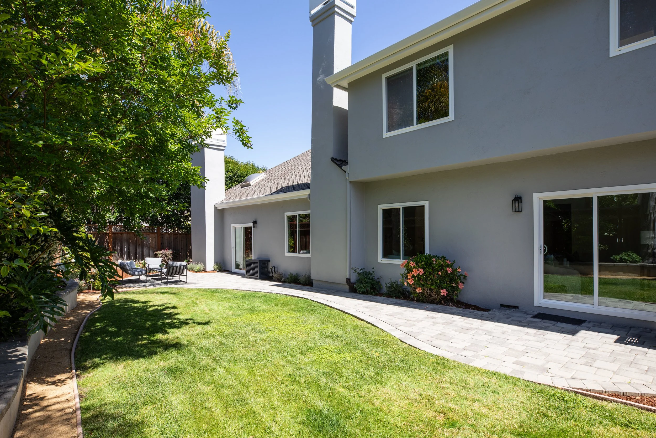 The backyard of a modern house with a gray exterior, large windows, and a small patio area. There is well-kept green grass, bushes, flowers, and a tree providing shade. Outdoor furniture is arranged on the patio.