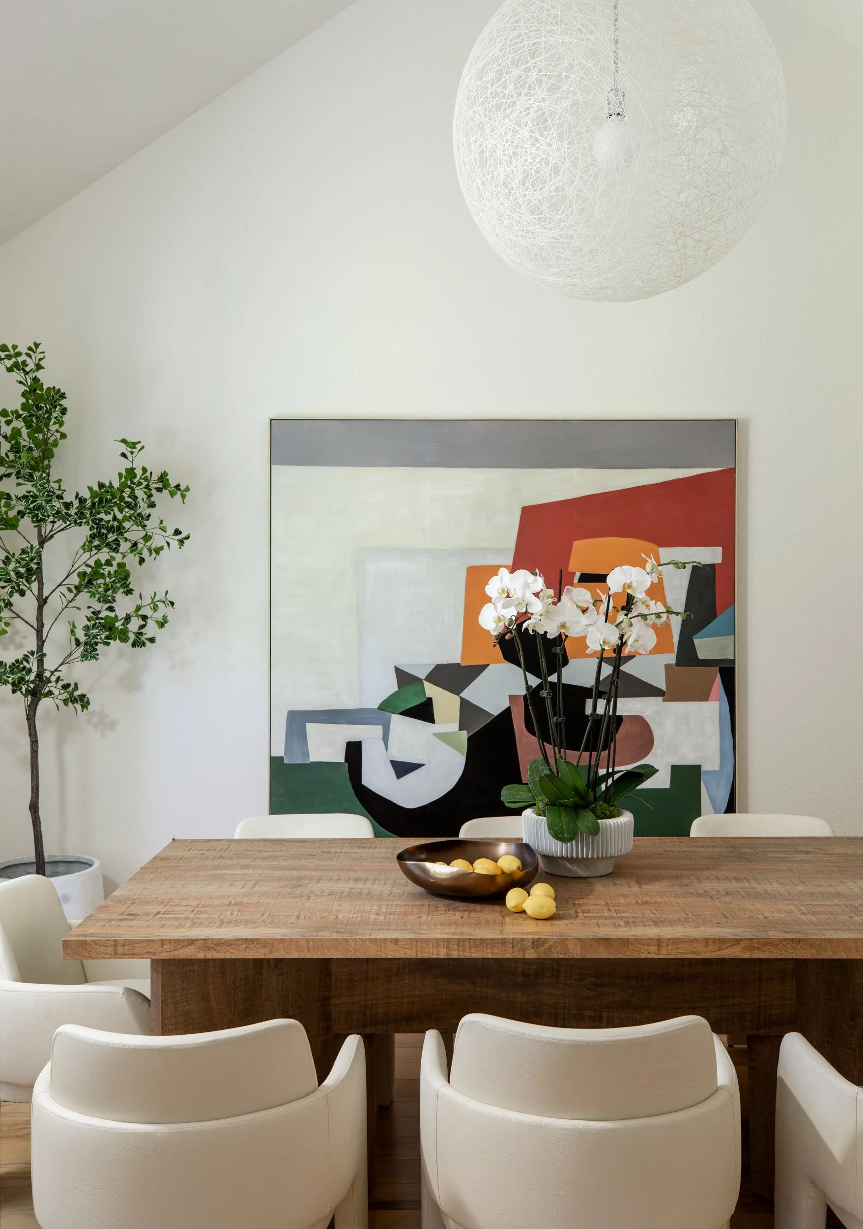 Modern dining room with a wooden table surrounded by six white chairs, decorated with a large potted orchid, a golden bowl with yellow lemons, an abstract painting on the wall, a potted tree in the corner, and a spherical white pendant light hanging 