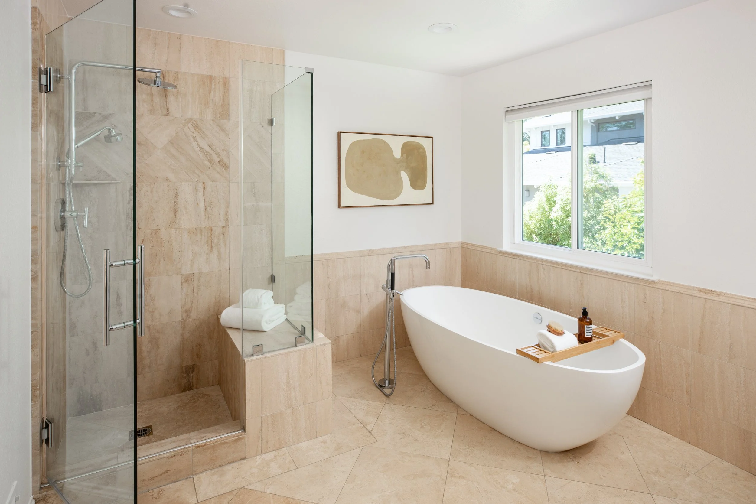 Modern bathroom with a walk-in shower with glass doors, a white freestanding bathtub with a wooden bath tray, a window letting in natural light, and beige tiled walls.