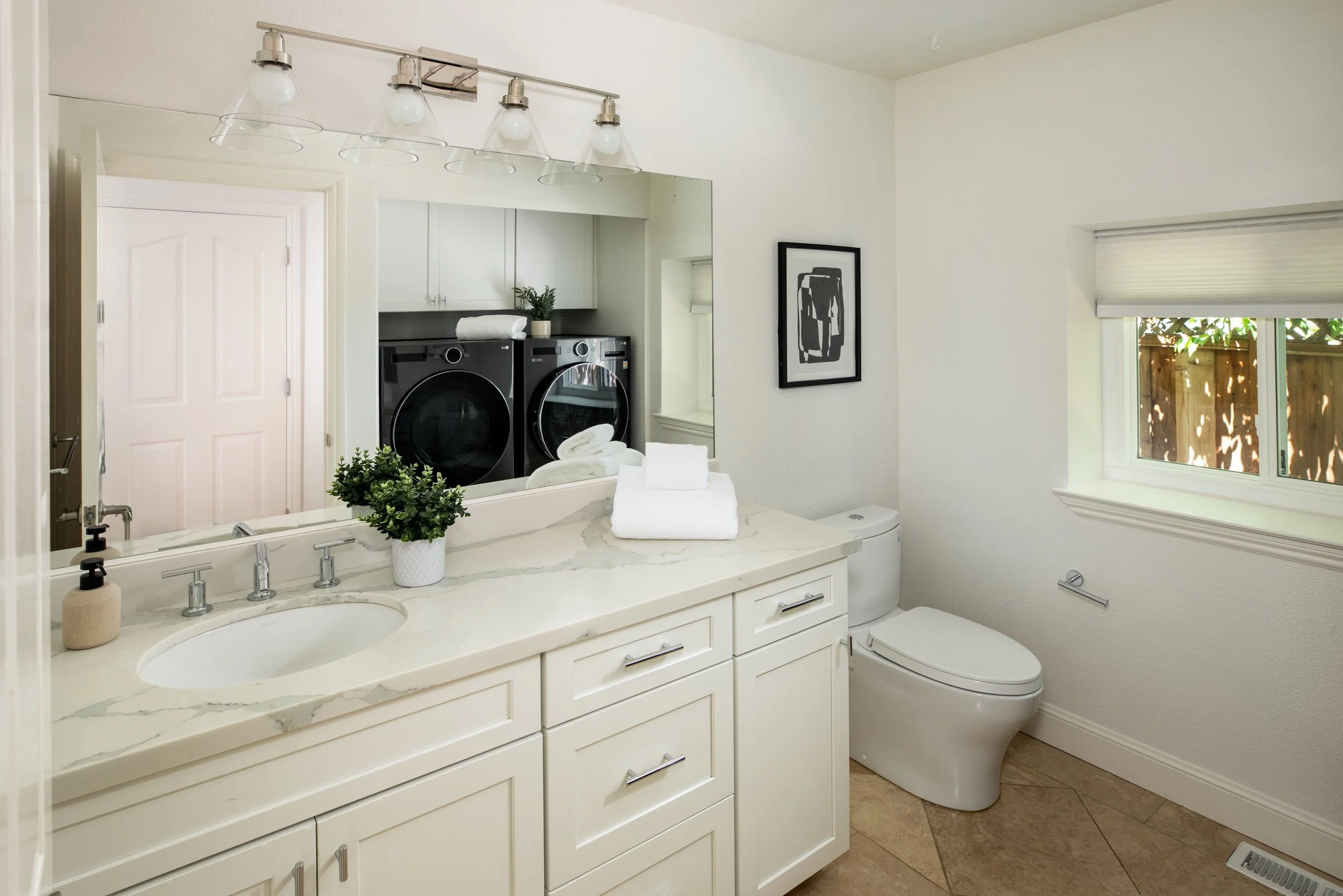 Bathroom with a white vanity, a marble countertop, a sink, a toothbrush holder, matching towels, a plant, a large mirror, a washer and dryer, a framed abstract art, a window with a blind, and a toilet.