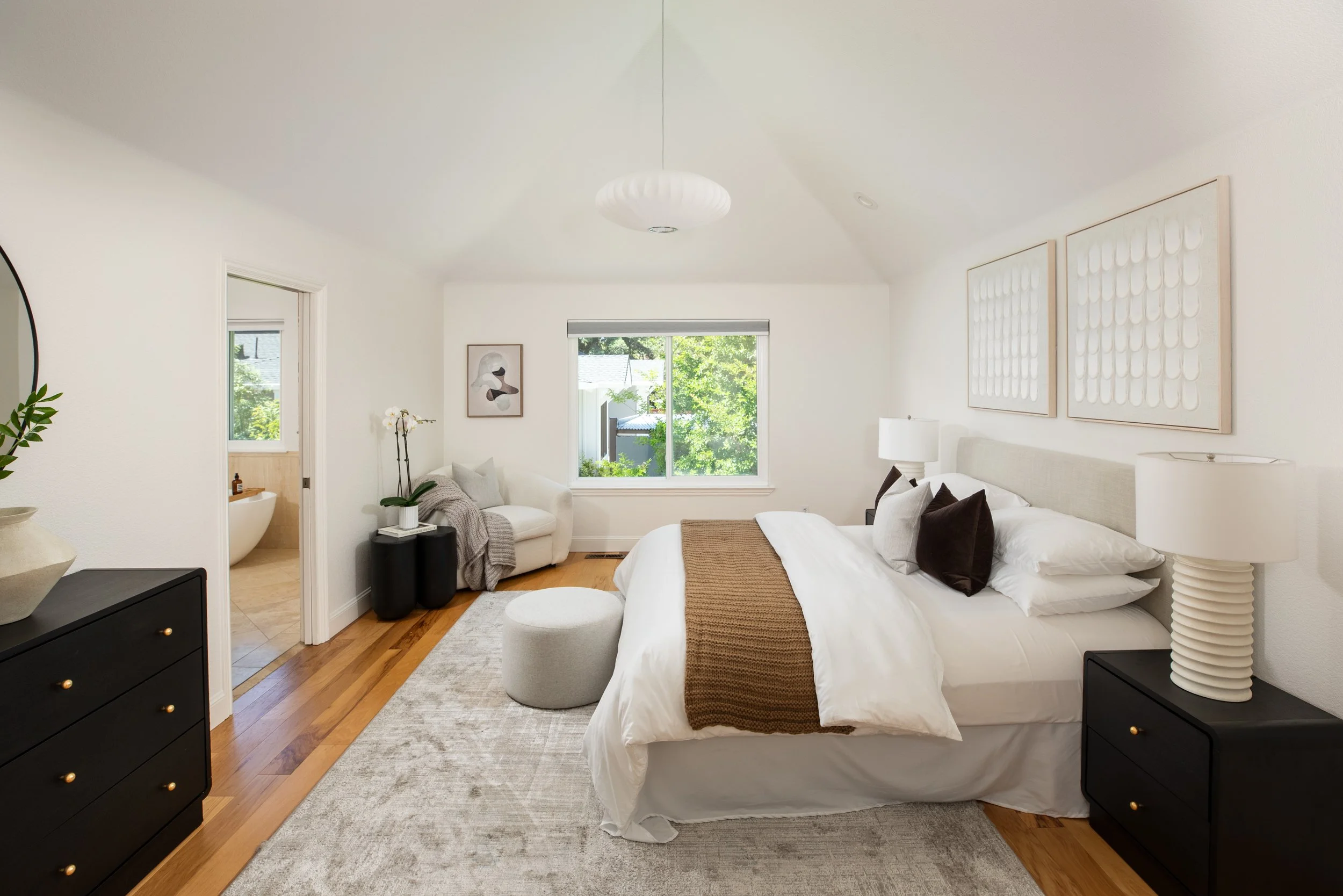 A modern bedroom with a large bed, black nightstands, artwork, a white armchair, a window showing greenery outside, and a view into a bathroom with a bathtub.