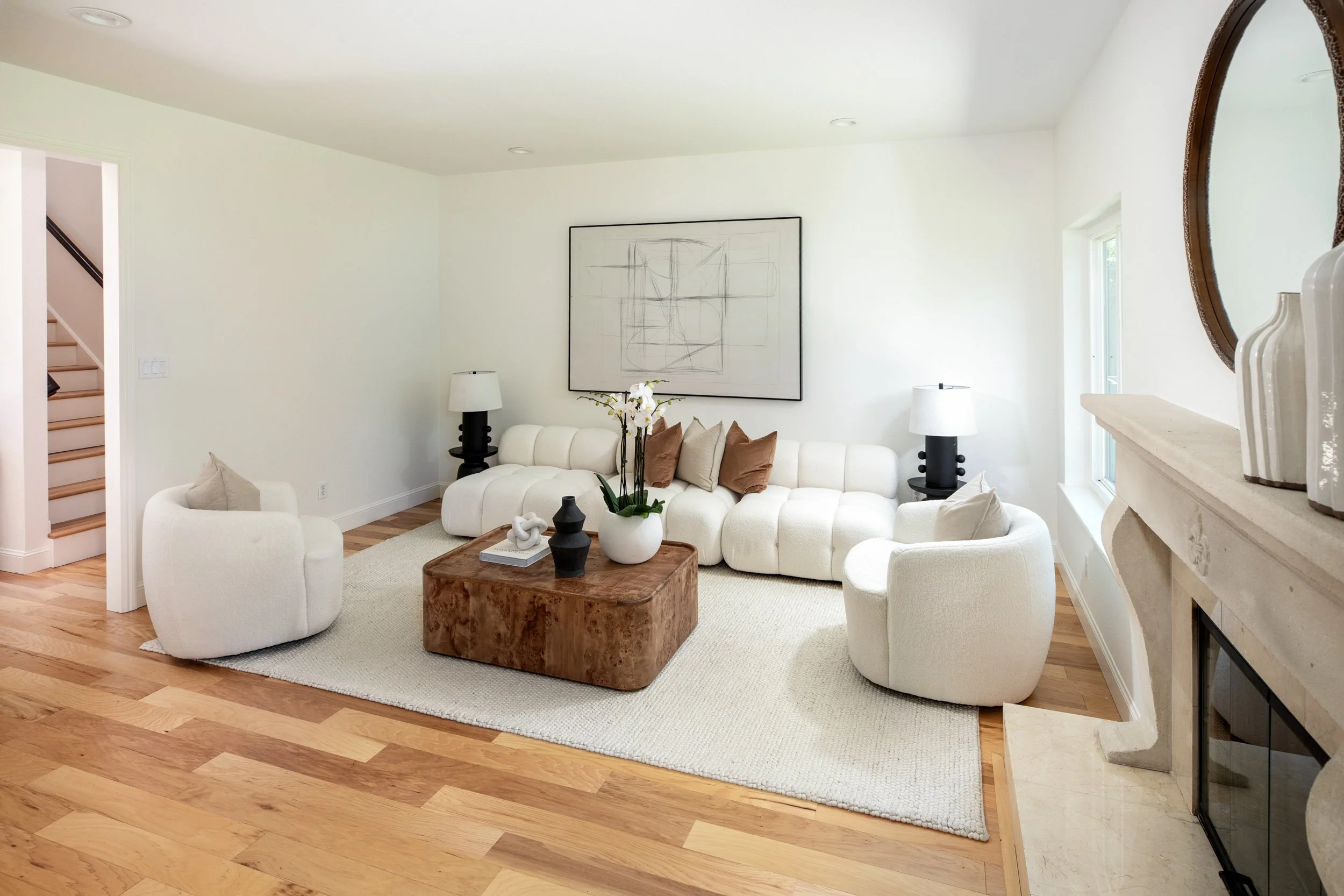 Living room with white sofa, armchair, wooden coffee table, black and white lamps, wall art, orchid plant, marble fireplace, and windows with natural light.