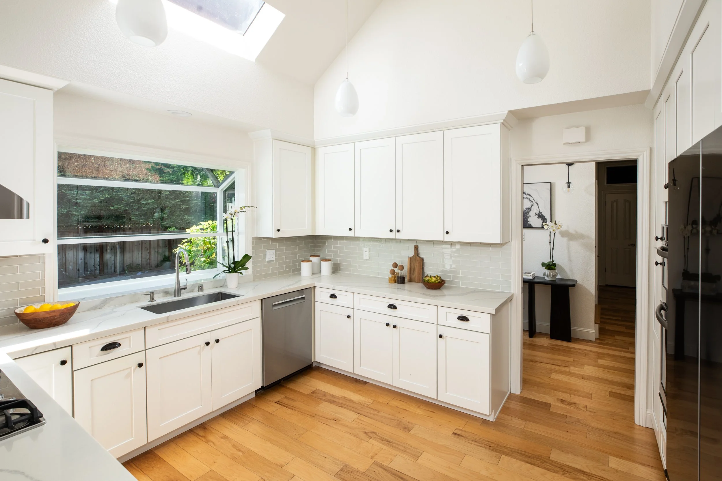 Bright kitchen with white cabinets, marble countertops, and a large window overlooking a backyard with trees. Decor includes a bowl of lemons, potted plants, and containers. Wooden flooring and modern light fixtures add to the clean, minimalist aesth