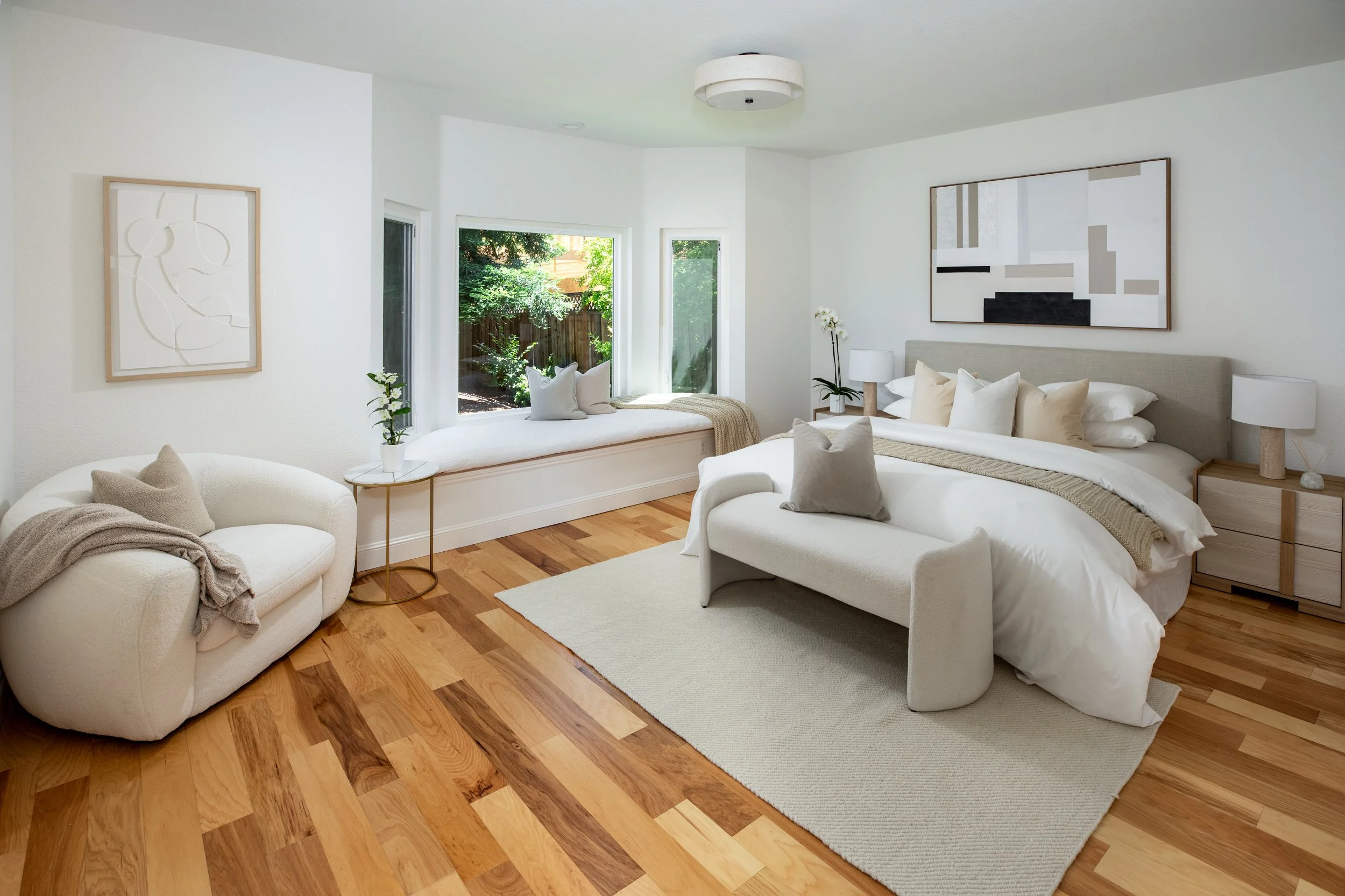 Brightly lit bedroom with white walls and wooden floors, featuring a bed with white linens and multiple pillows, a side table with lamps, a window seat with cushions, large abstract artwork on the walls, and large windows showing greenery outside.