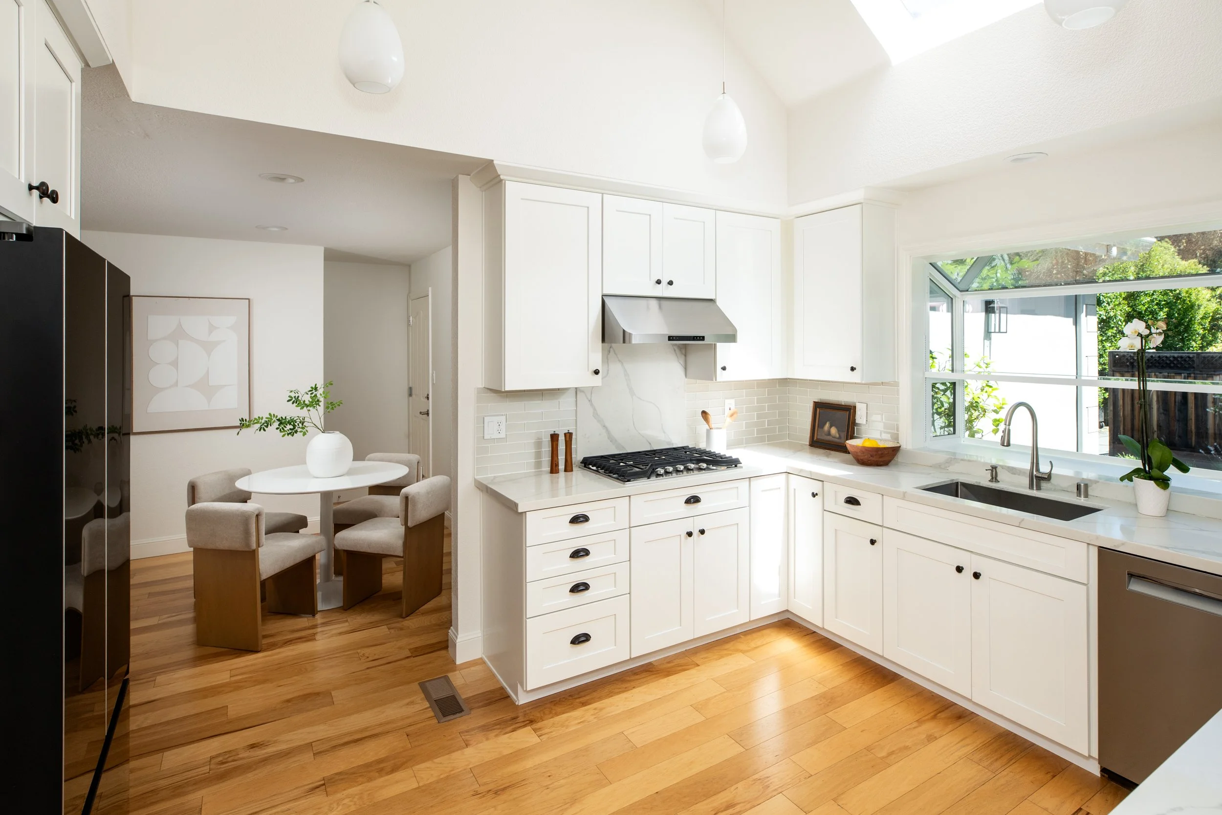 A bright, modern kitchen with white cabinetry, a marble backsplash, and wooden flooring. There is a window above the sink allowing natural light, and a small dining table with beige chairs on the left side of the image.