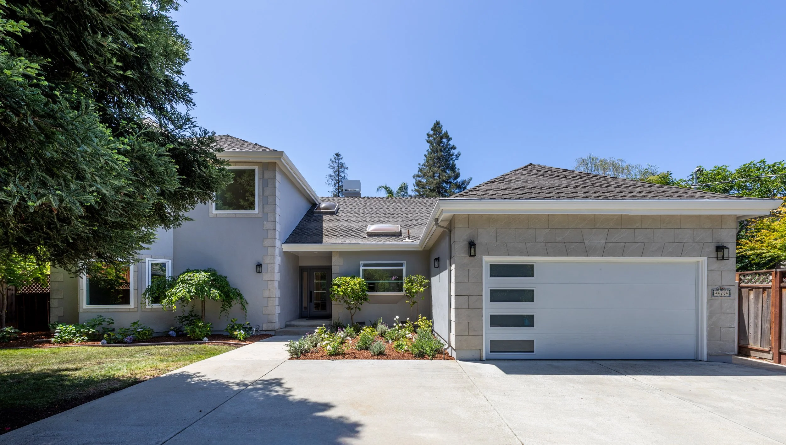 Front view of a modern house with a gray stone exterior, a white garage door with rectangular windows, a front door, well-maintained lawn, shrubs, and trees, under a clear blue sky.