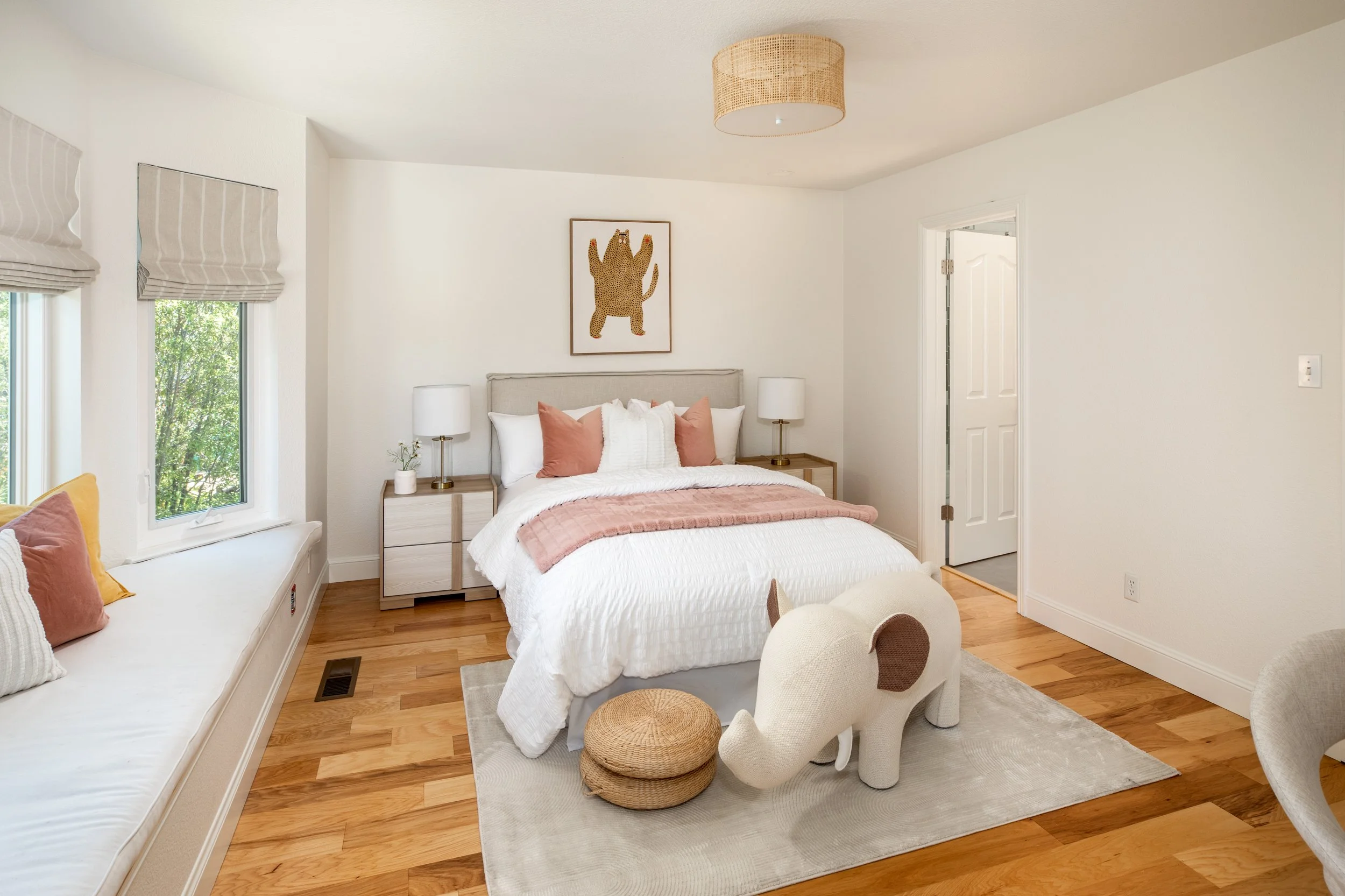 A bedroom with white walls, a bed with white bedding and pink pillows, two nightstands with white lamps, a framed picture of a bear on the wall, windows with striped shades, a built-in window seat with pillows, and a white armchair with a cushion, on