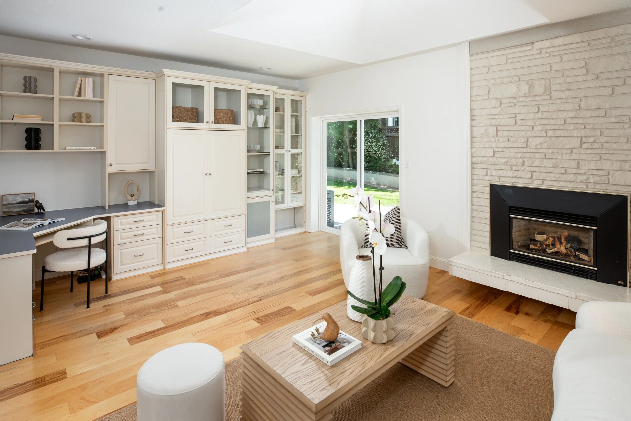 Bright living room with white built-in shelves, a glass door leading outside, a stone fireplace with a fire, and a wooden coffee table with decorative items.