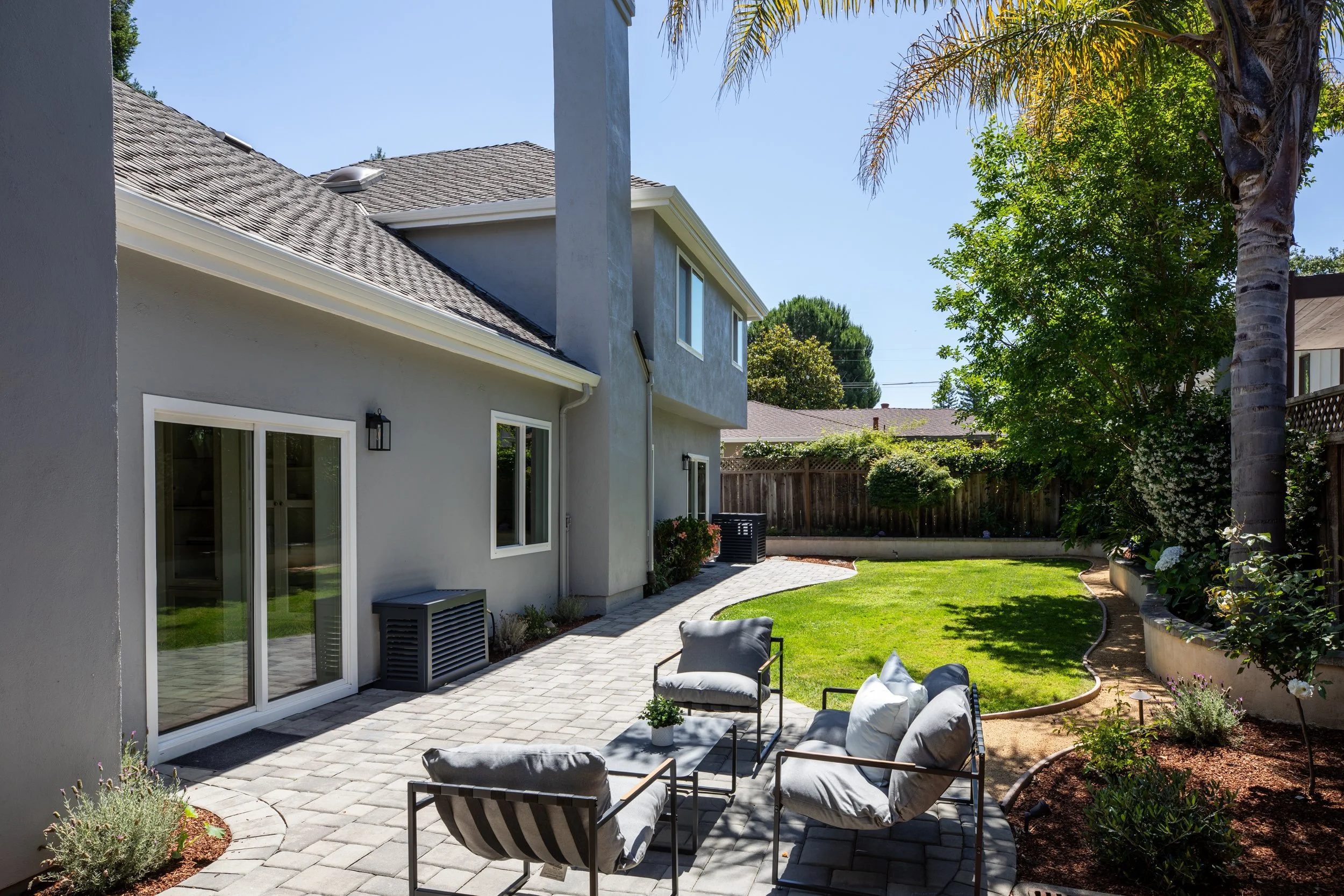 Backyard patio with outdoor furniture, potted plants, a grassy lawn, and a house with sliding glass doors and windows surrounded by trees and a wooden fence.