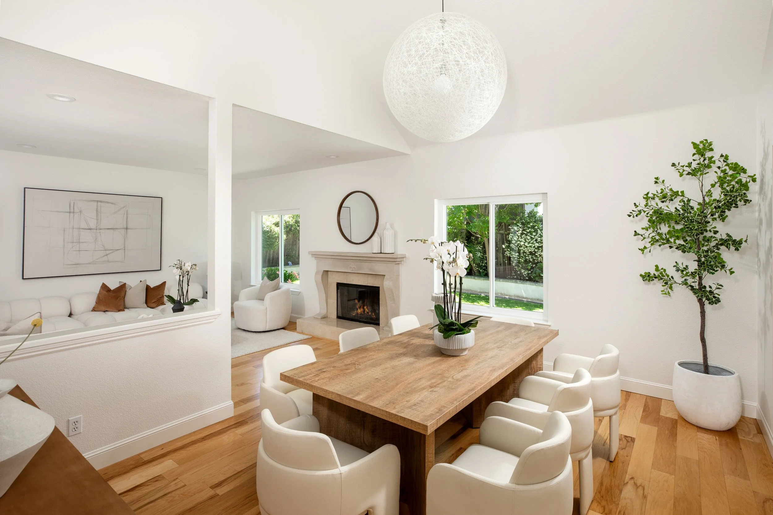 A bright living and dining room with white walls, hardwood floors, and large windows. There is a wooden dining table with white chairs, a white sofa with brown pillows, a white armchair near a fireplace, and a large potted tree to the right. A spheri