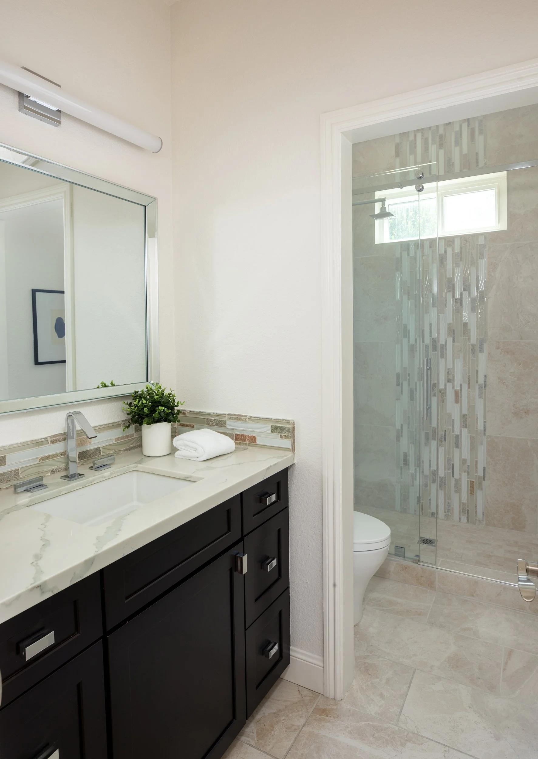 Modern bathroom with black vanity, white countertop, large mirror, plant, rolled towel, and walk-in shower with glass door and tile decor.