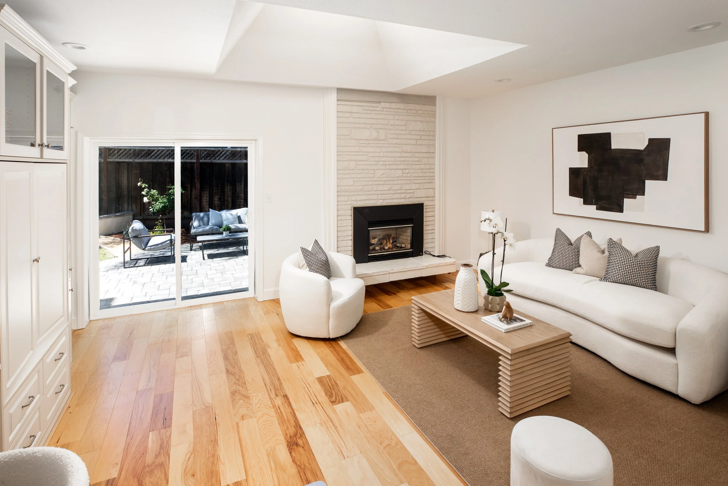 Living room with cream-colored furniture, artwork, and hardwood floors, leading to an outdoor patio with seating and a garden area.
