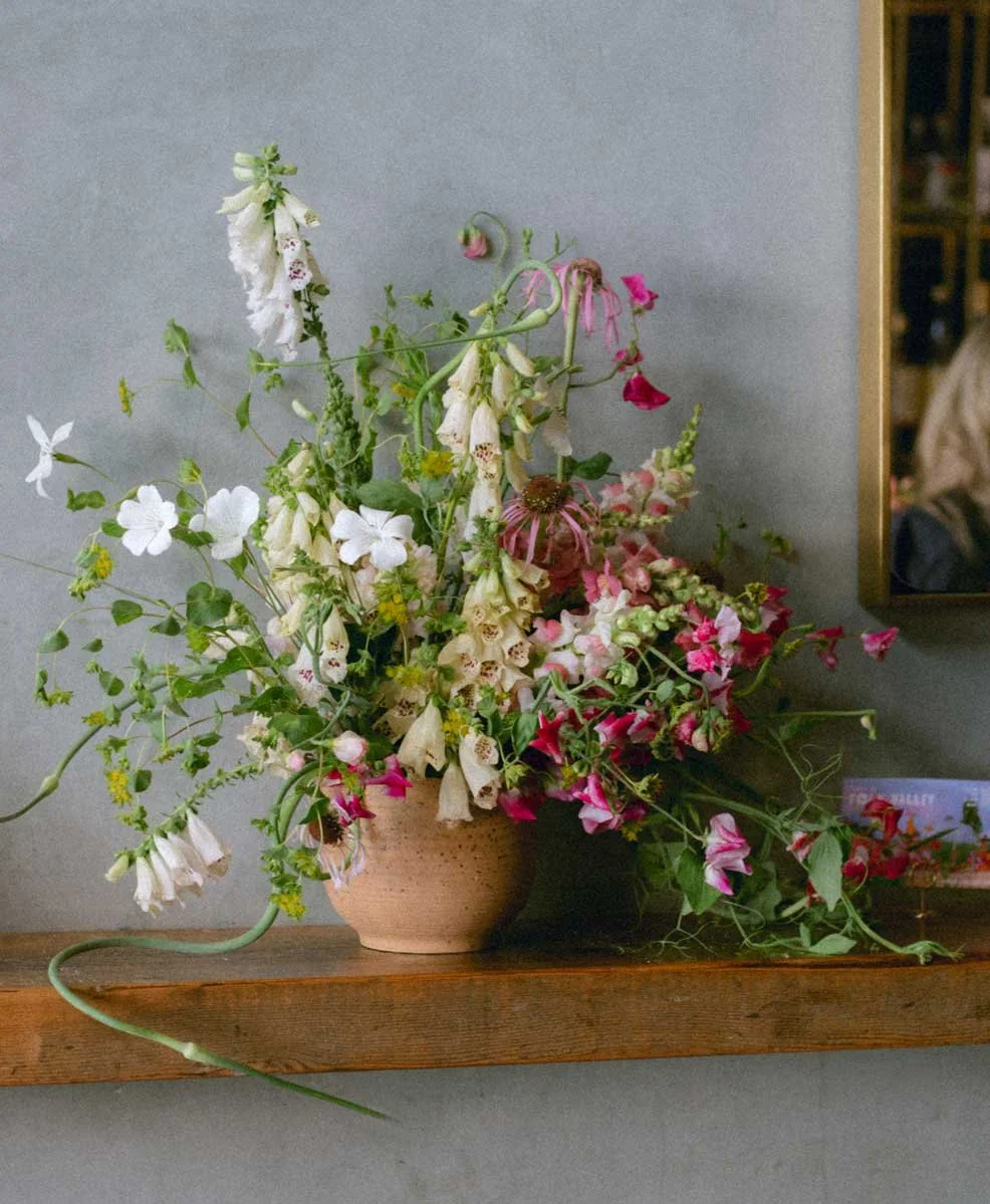 A floral arrangement with white and pink flowers in a rustic ceramic vase on a wooden shelf against a light gray wall.
